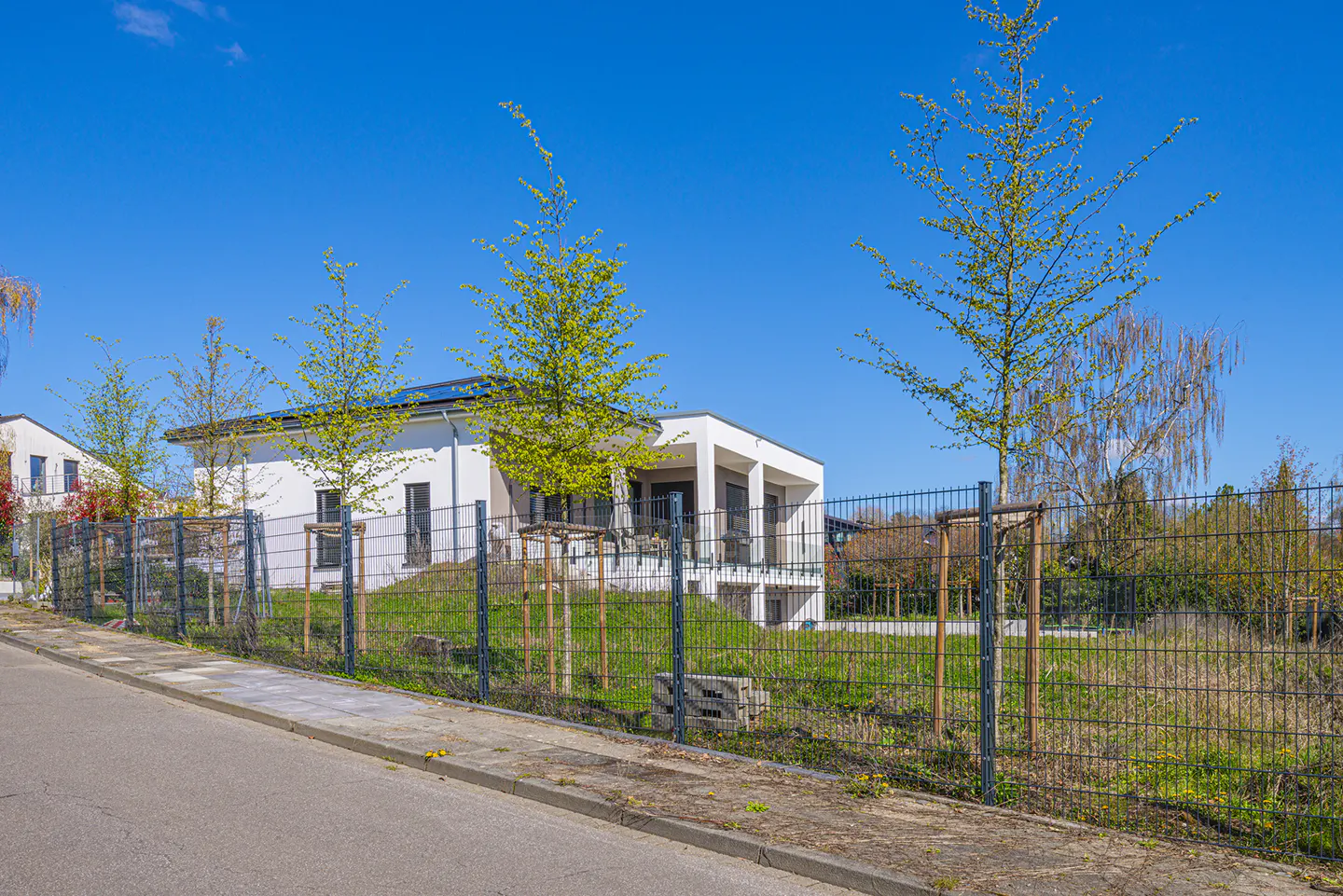 A modern white house with a gray fence and green trees under a clear blue sky.