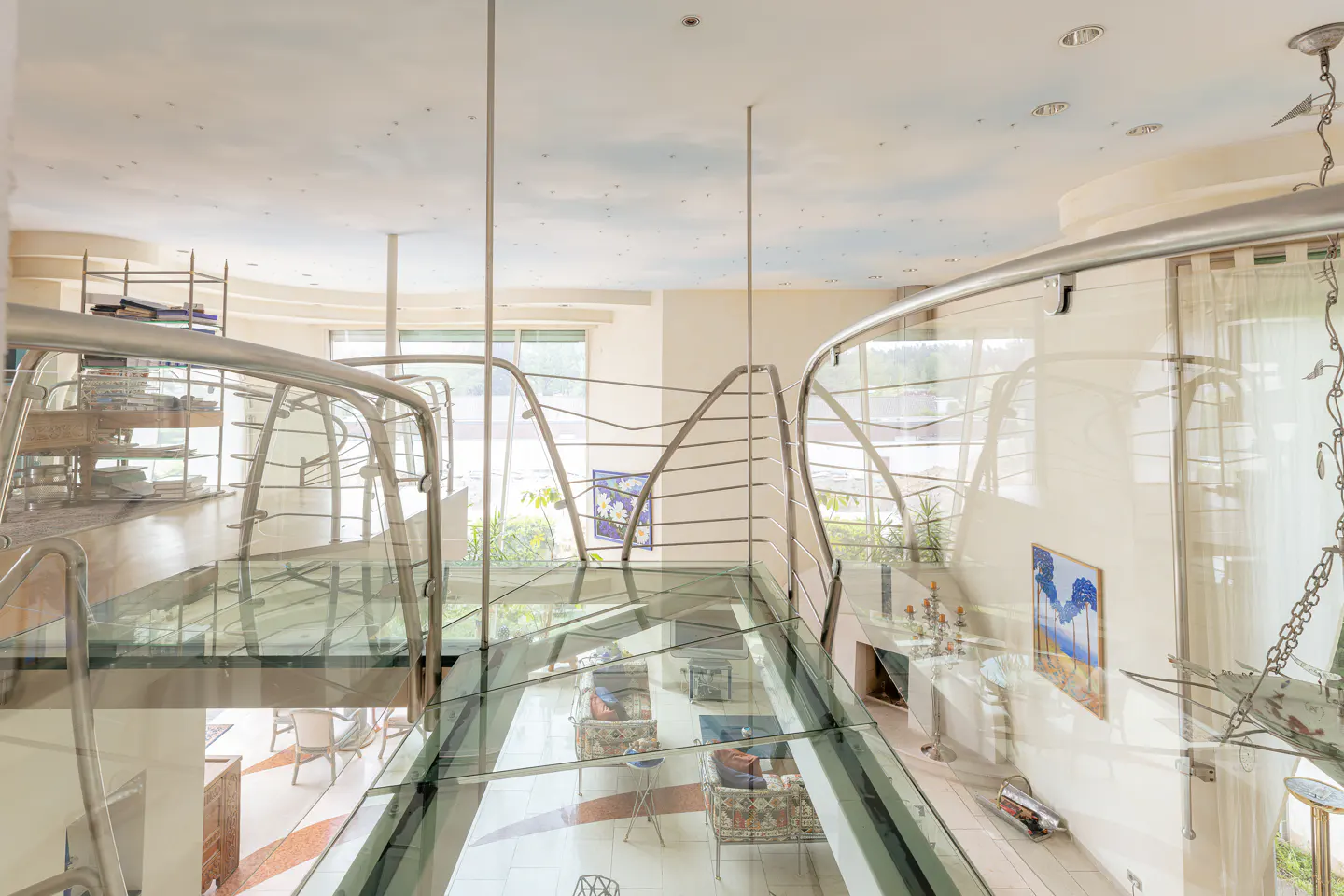 A glass walkway with metal railings overlooks a bright living room with a painted sky ceiling and modern decor.