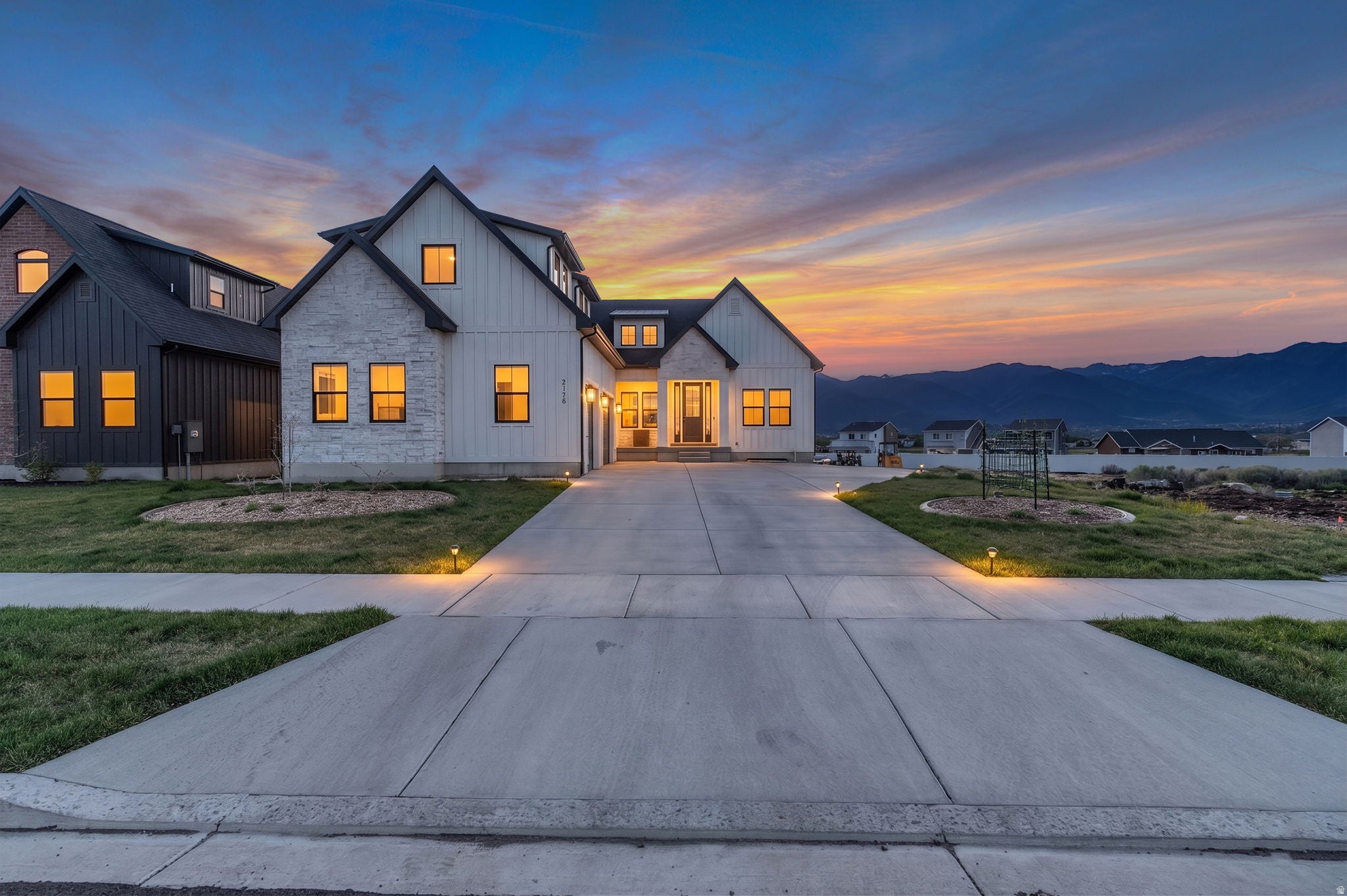 Modern white farmhouse with stone accents and glowing windows at sunset, featuring a concrete driveway and mountain backdrop. [AI-generated text]