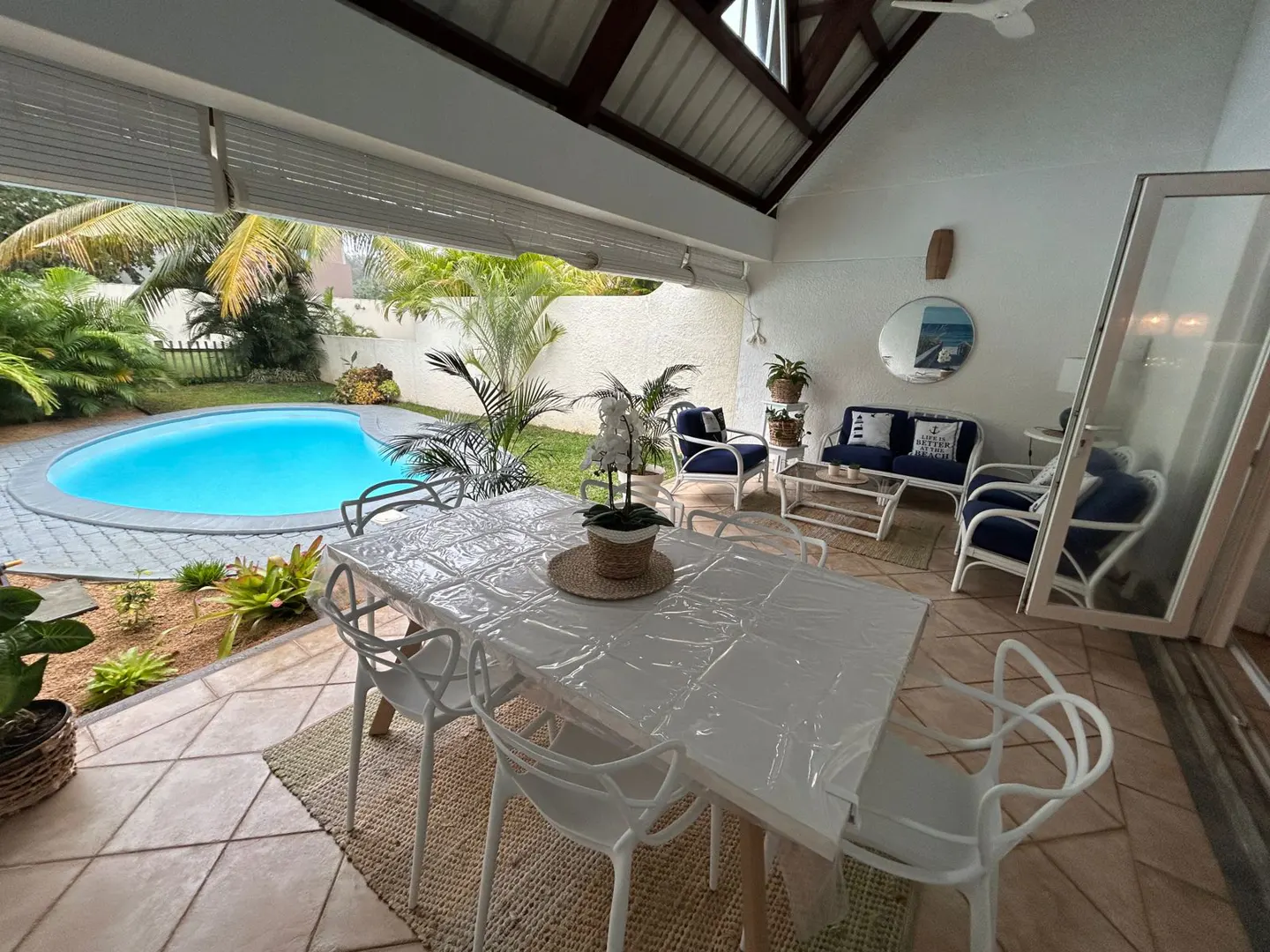Outdoor patio with a white table, chairs, and a view of a blue pool surrounded by lush greenery. Seating area with blue cushions in the background.