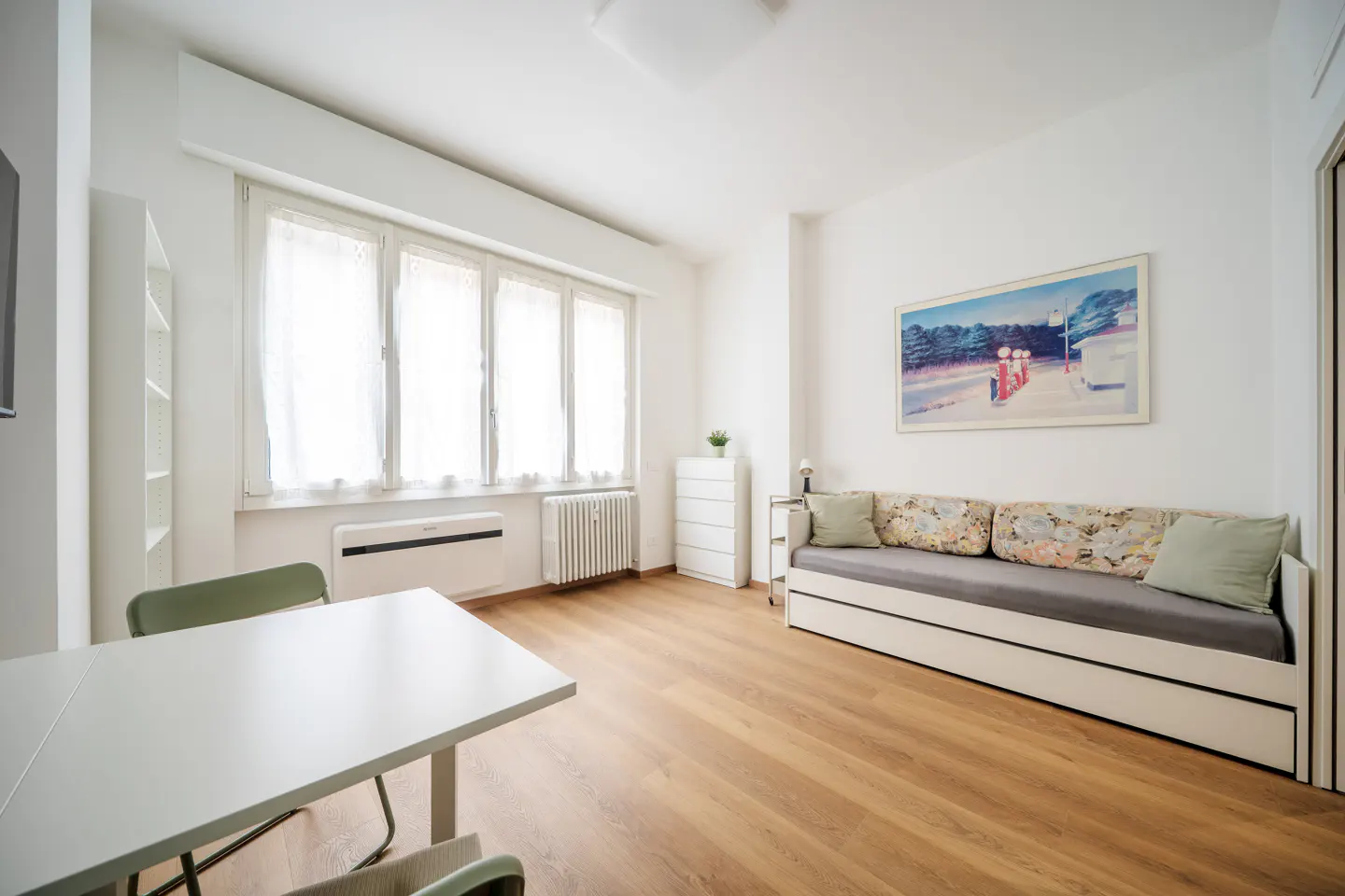 Bright, modern apartment interior with a white table, green chairs, and a daybed with floral pillows. Hardwood floors and natural light.