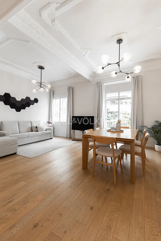 Bright, open-plan living and dining area with light wood floors, white walls, and modern lighting fixtures. A gray sofa sits near a black geometric wall decoration.