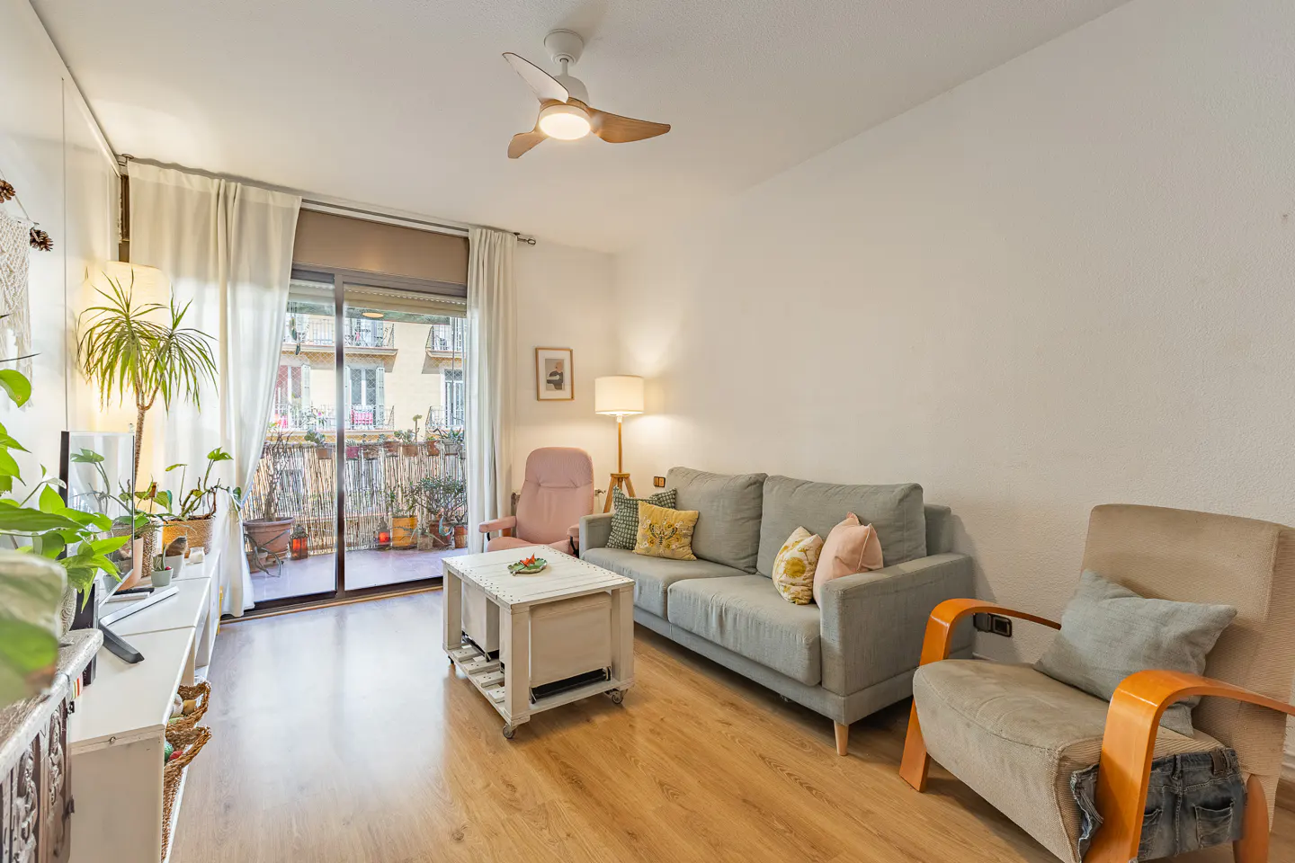 Bright living room with a gray sofa, beige armchair, and a balcony filled with plants. A white ceiling fan is visible.