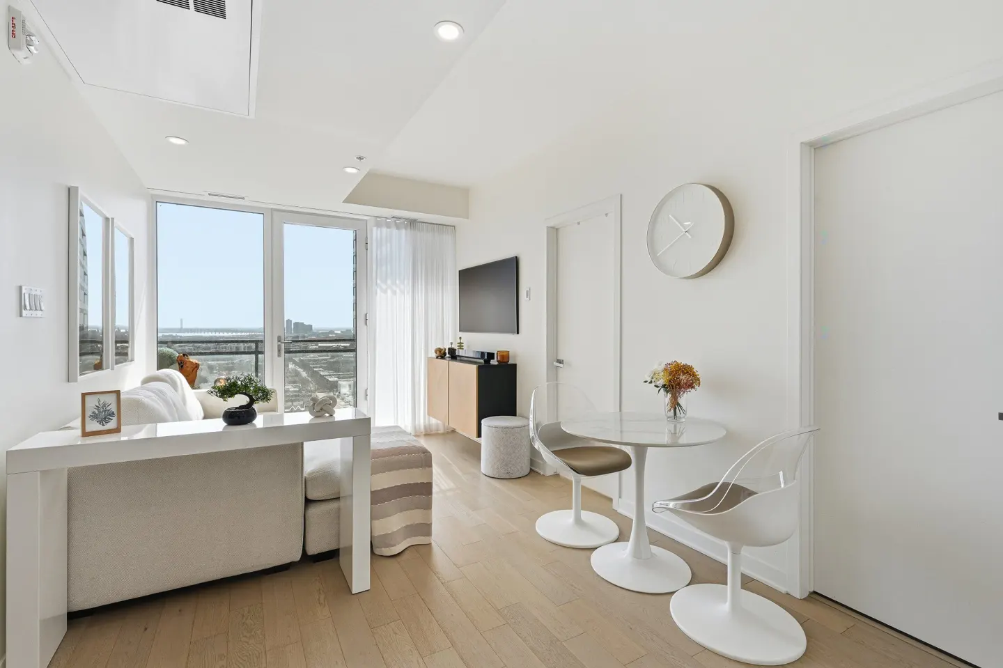 Bright, modern condo interior with white walls, light wood floors, and a balcony view. A white table sits behind a sofa, and a round dining table with chairs is nearby.