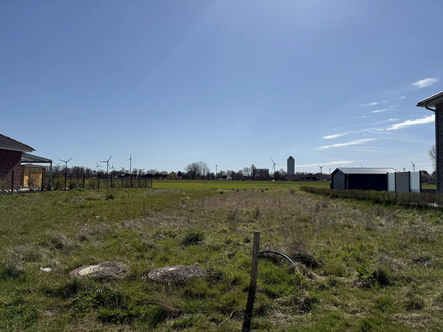 Vacant lot with green grass under a blue sky. Wind turbines and a tall building are in the background.