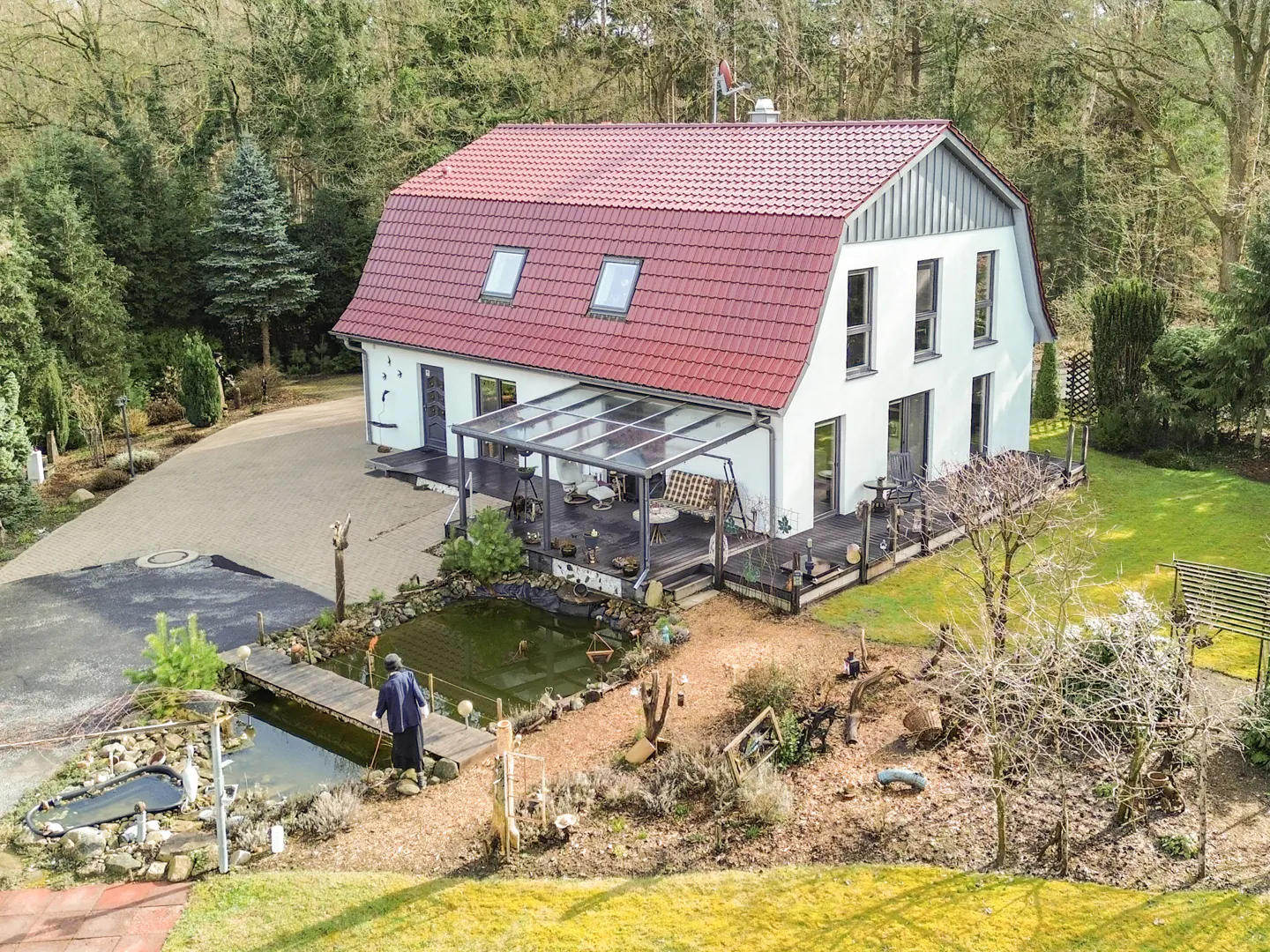 Aerial view of a white two-story house with a red roof, a pond, and a person standing on a small wooden bridge.
