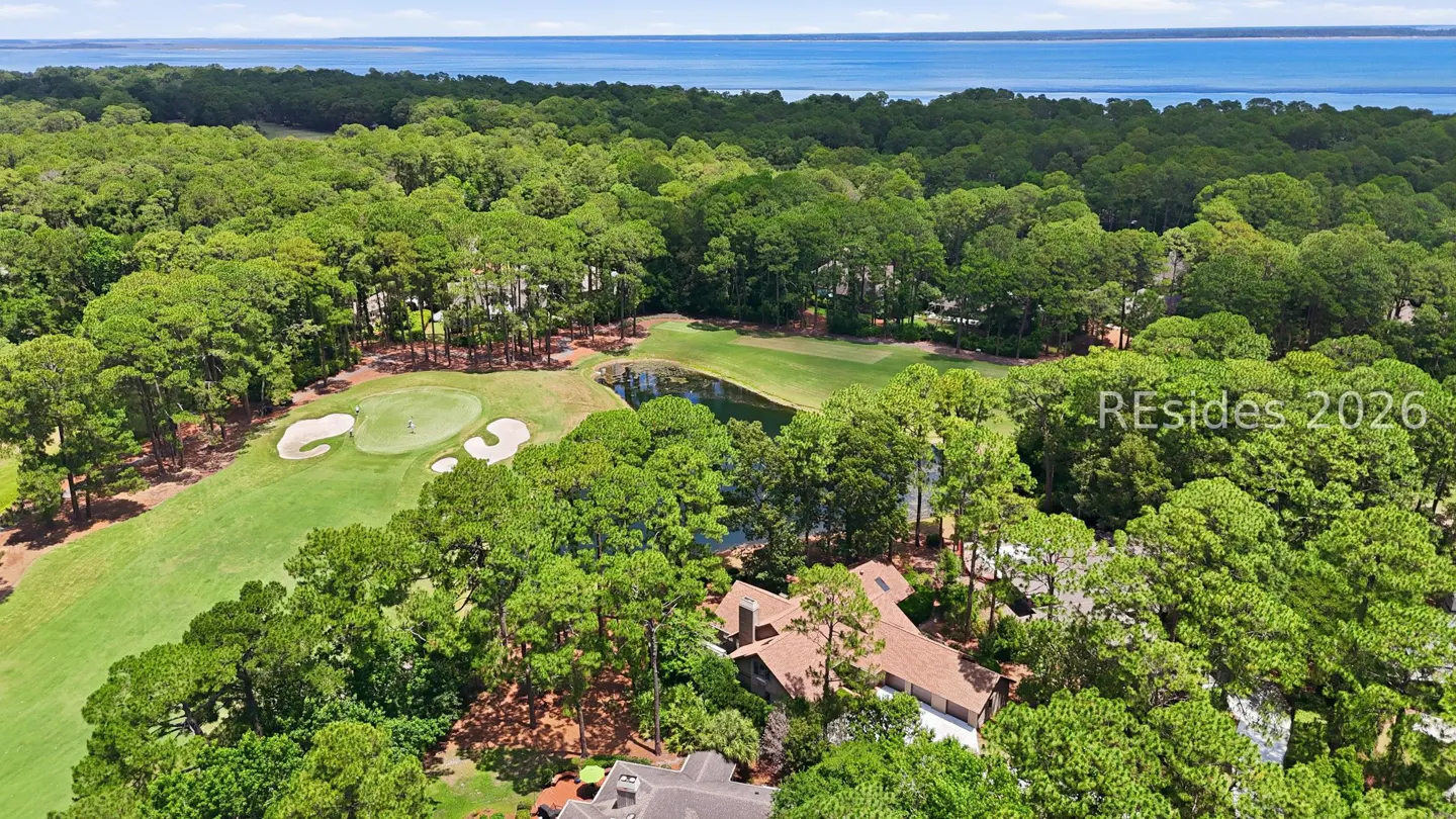 Aerial view of a house nestled in a lush forest next to a golf course green, a pond, and a distant blue ocean horizon.