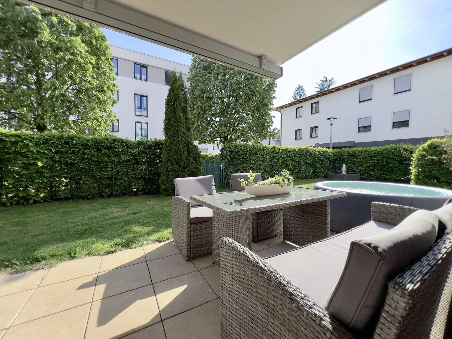 Outdoor patio with wicker furniture, a table with a centerpiece, and a hot tub, all set against a green lawn and hedge.