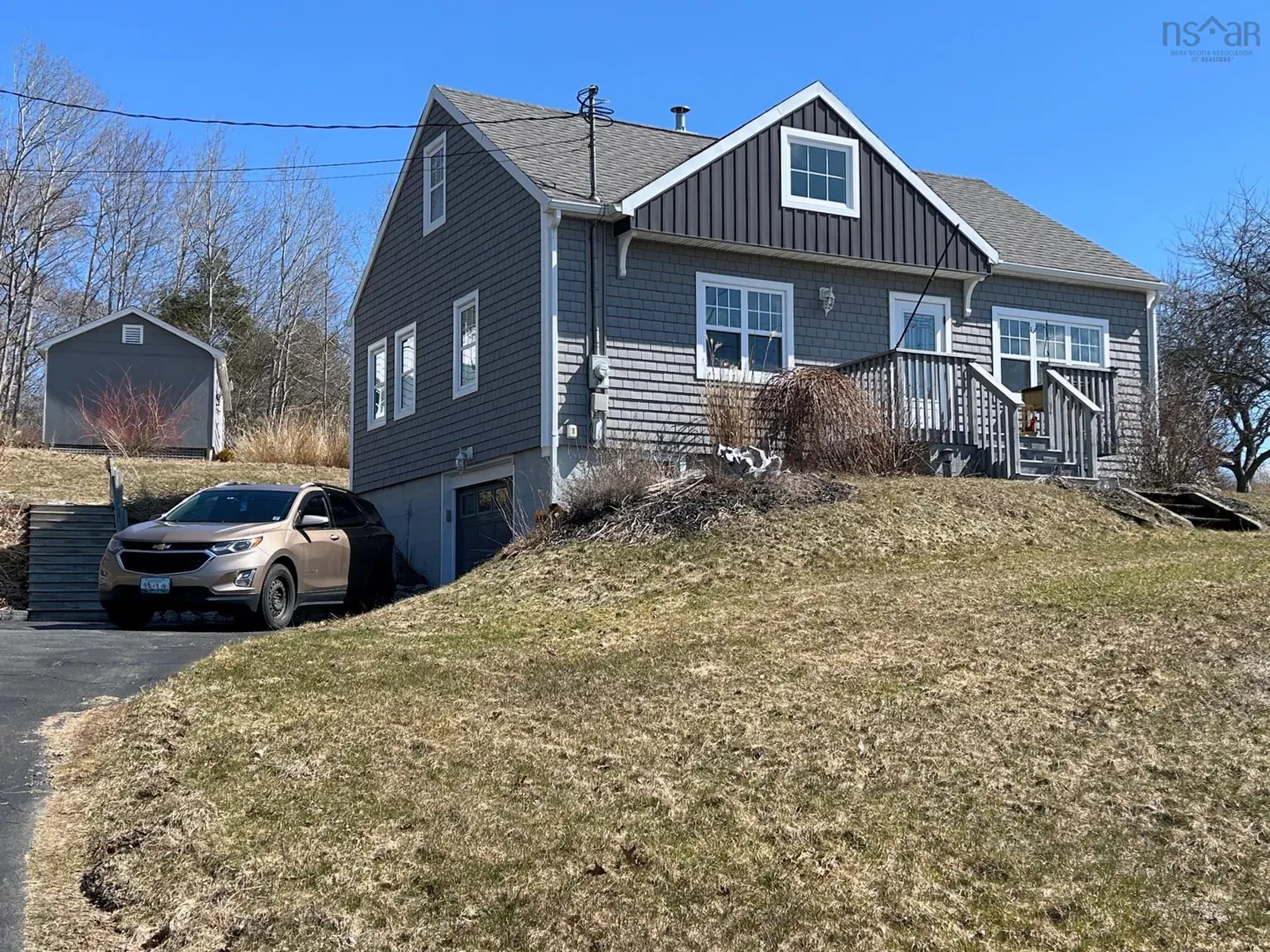 A gray two-story house with white trim sits on a grassy hill under a blue sky. A brown SUV is parked in front of a gray shed.