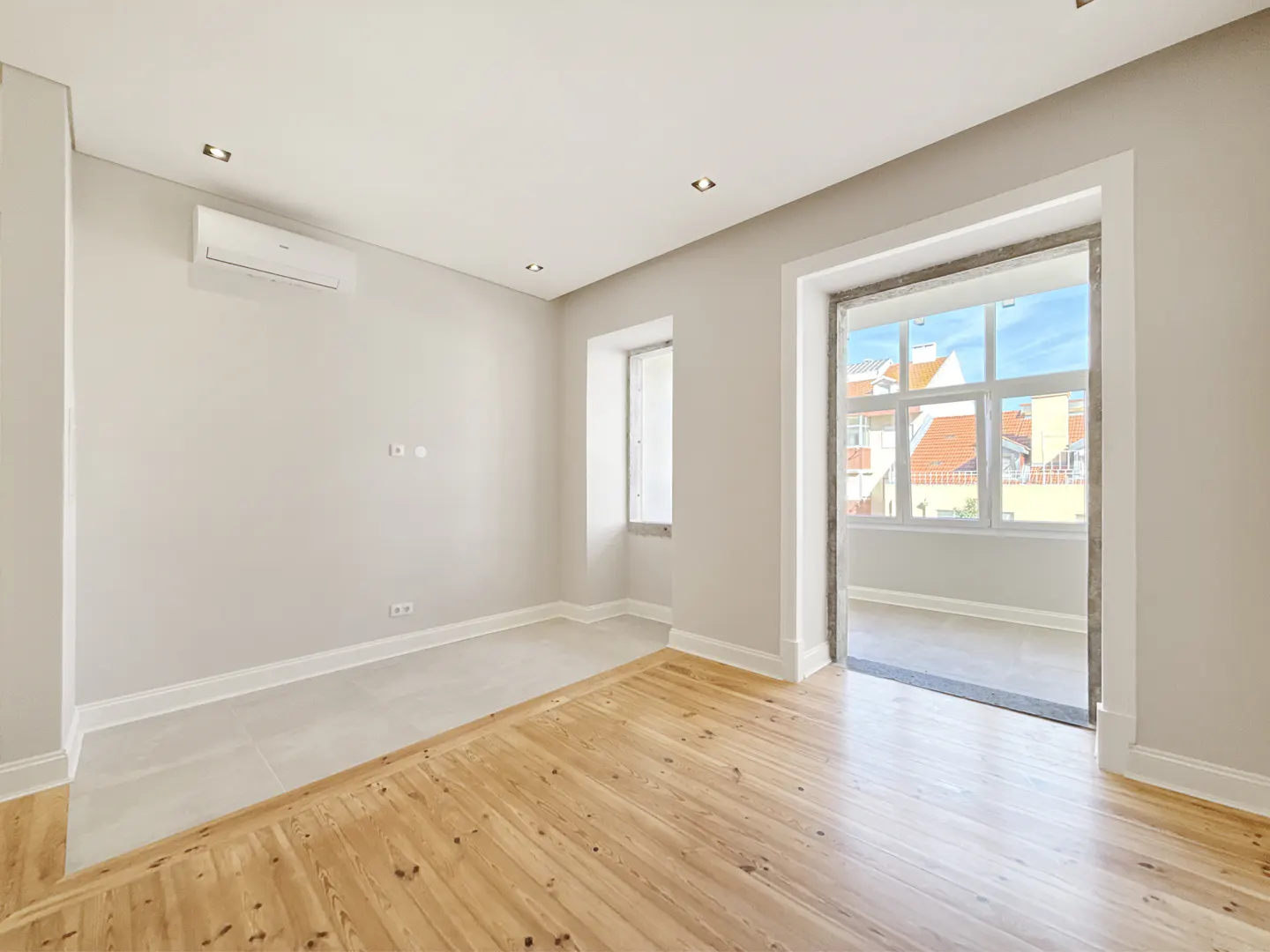 An empty room with light gray walls, wood floors, and a balcony view. A white air conditioner is mounted on the wall.