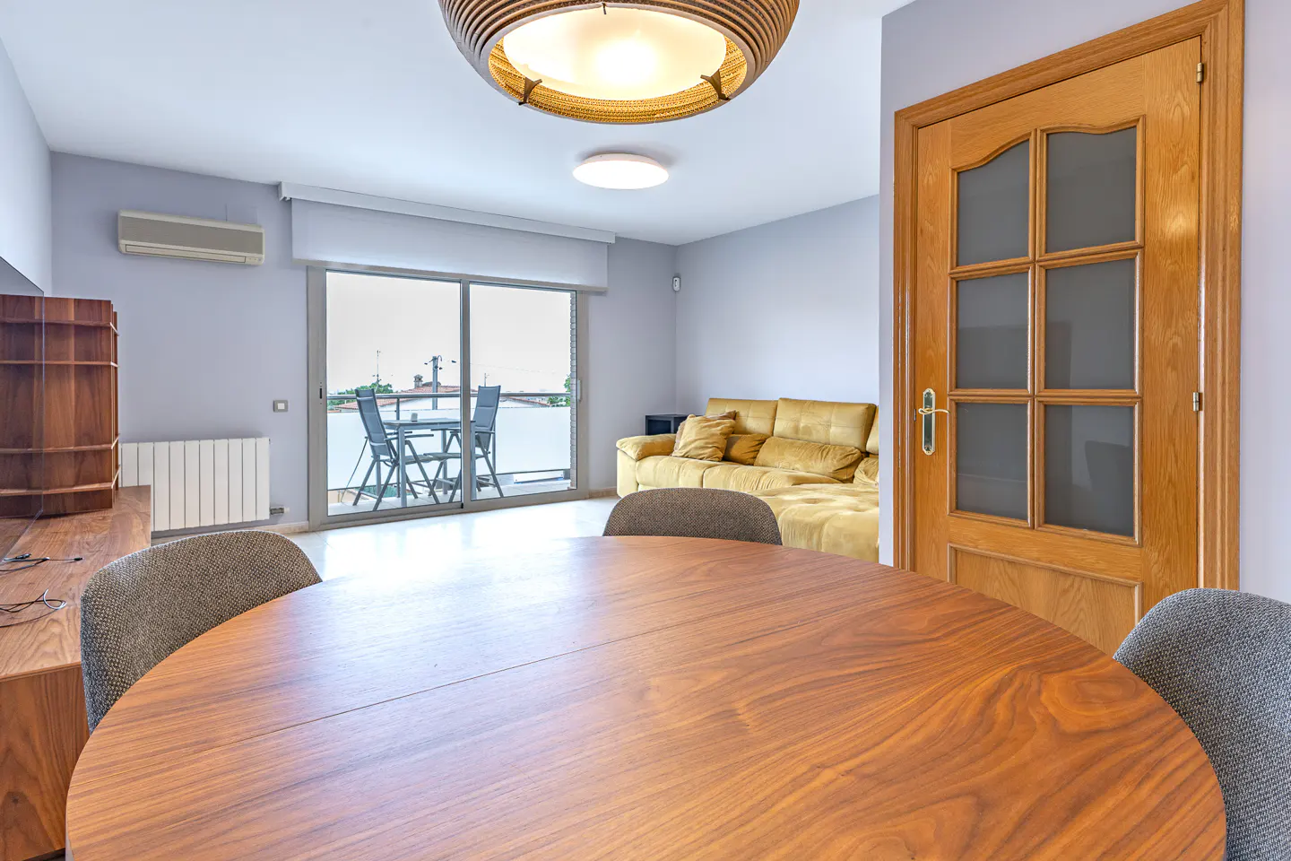 A bright living room with a wooden table, gray chairs, and a yellow sofa. A balcony with a table and chairs is visible through sliding glass doors.
