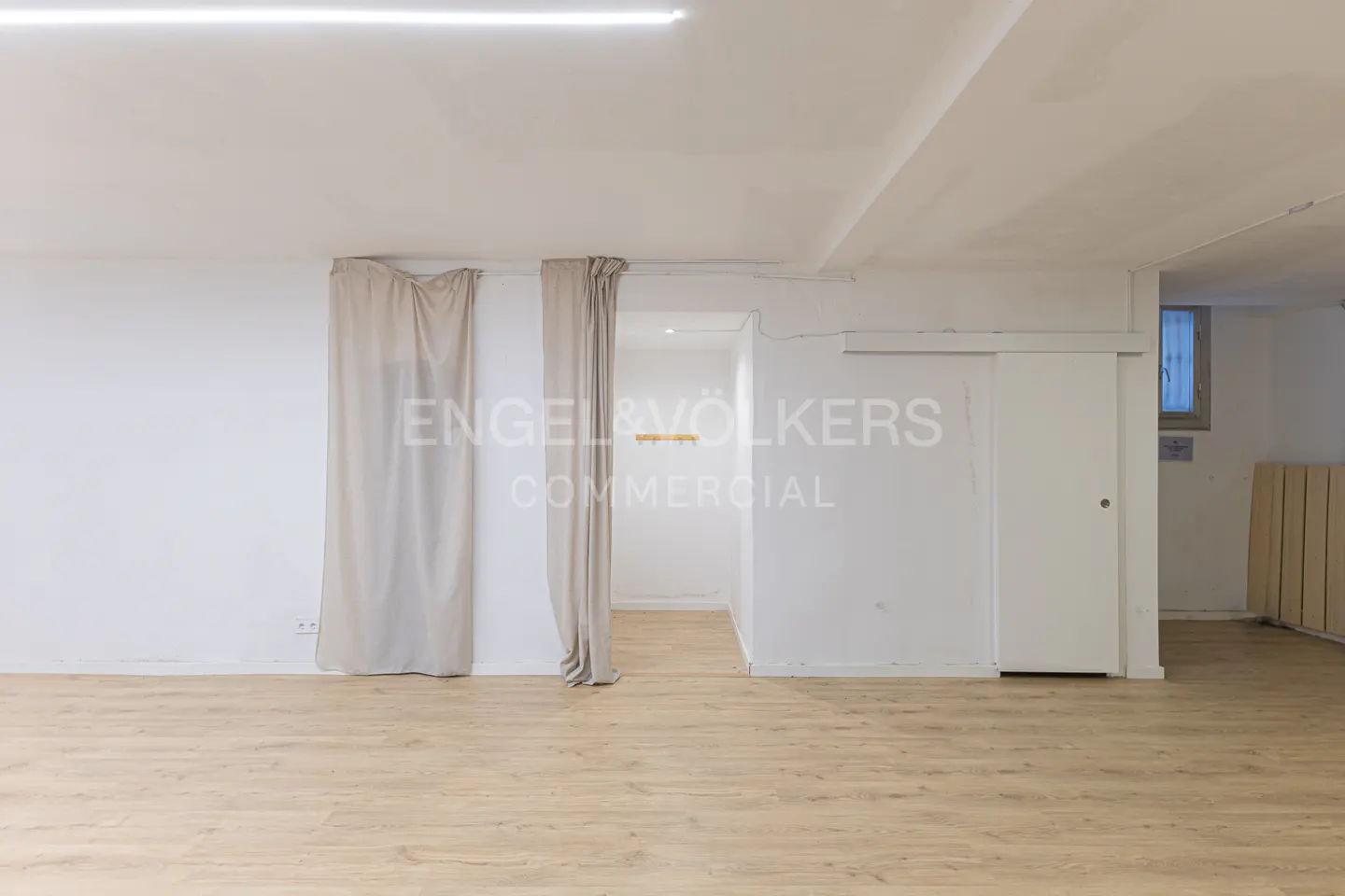 Empty room with light wood floors, white walls, and beige curtains. Engel & Völkers Commercial logo on the wall.