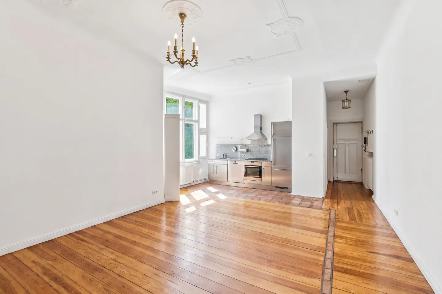 Bright open-plan room with hardwood floors, a modern stainless steel kitchen, and a classic chandelier.
