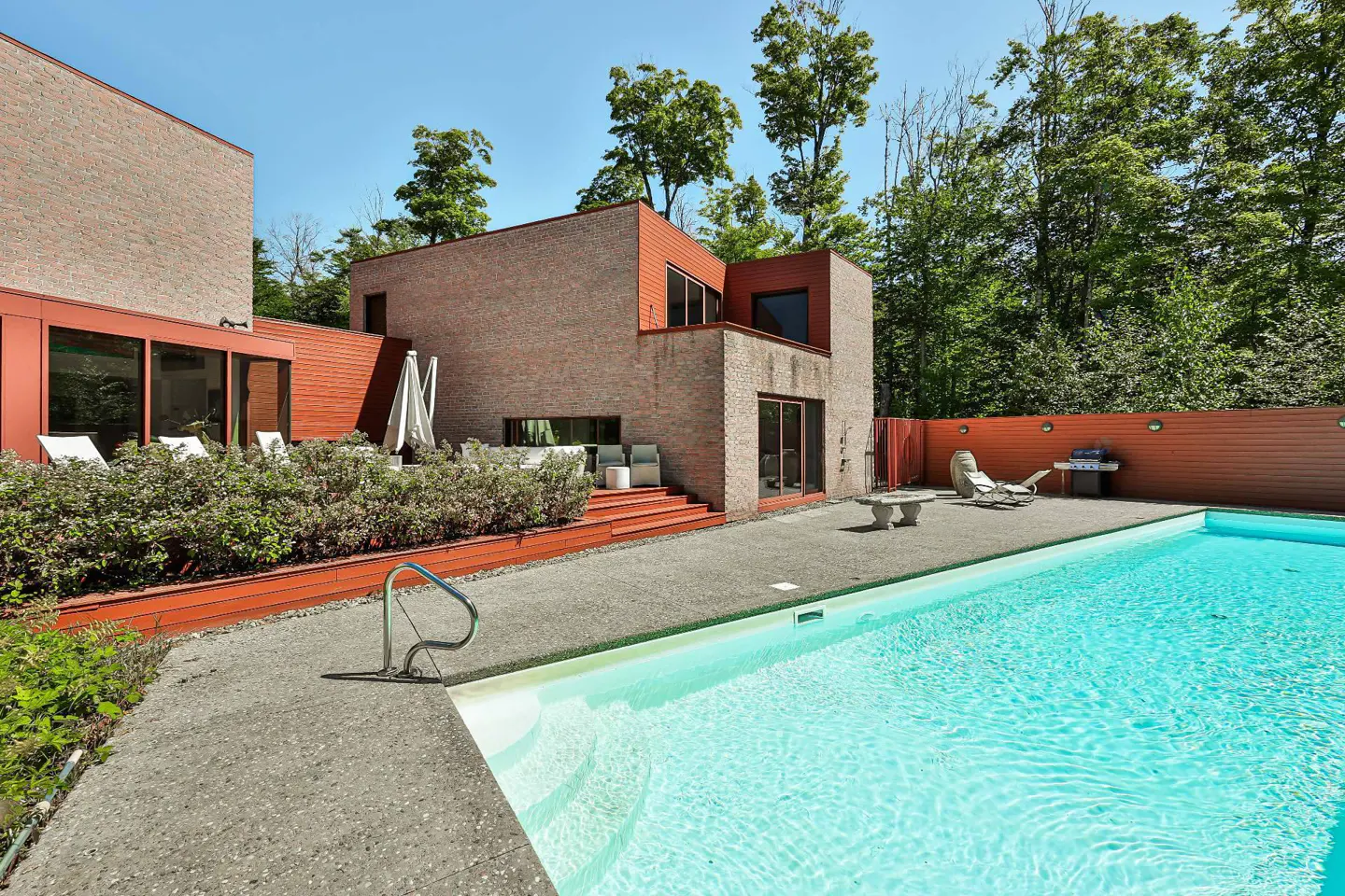 Exterior view of a modern brick house with a pool, lounge chairs, and a grill on a sunny day.