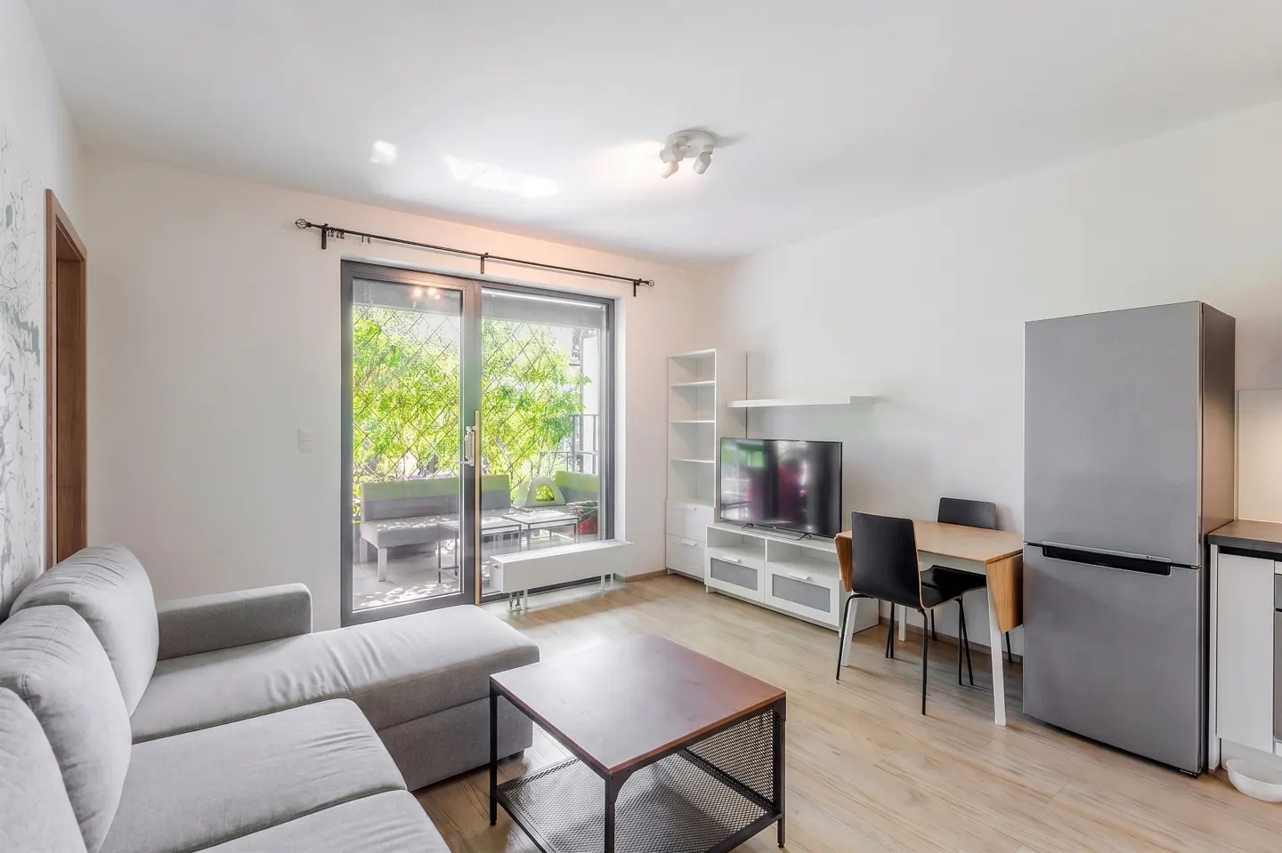 Modern studio apartment with a grey sectional sofa, coffee table, TV, dining set, and a stainless steel refrigerator.