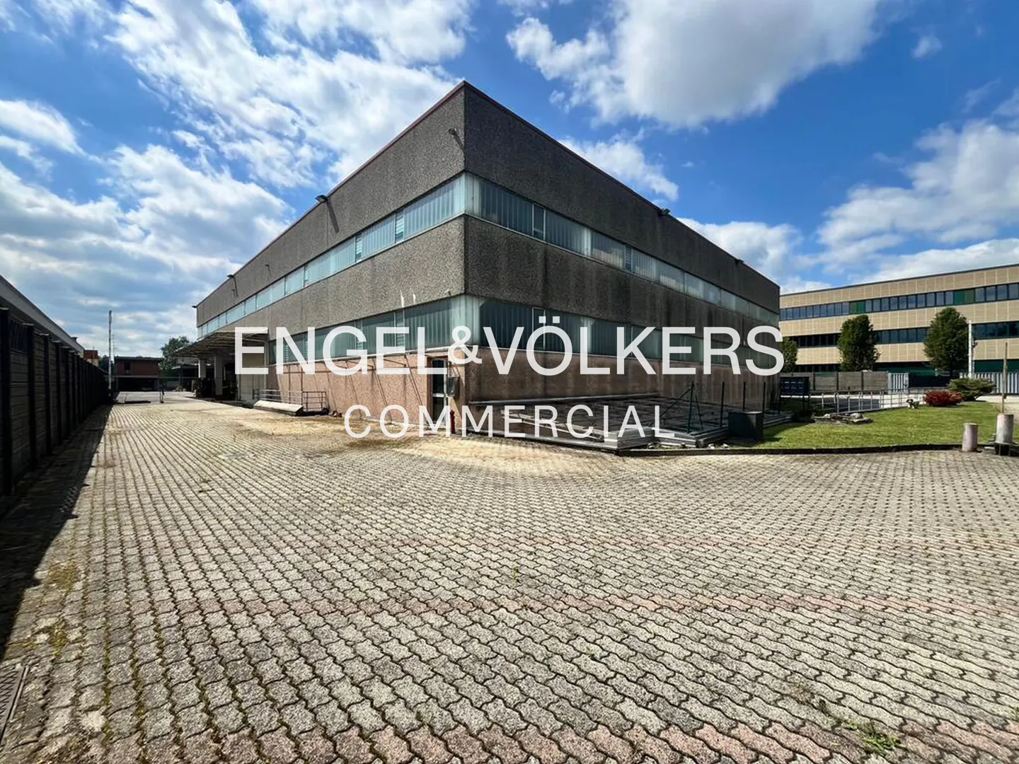 Exterior view of a gray commercial building with a brick parking lot under a blue sky. Engel & Völkers Commercial logo overlay.