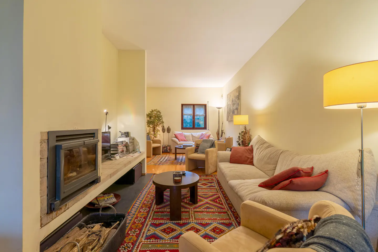 Living room with a fireplace, beige sofa with red pillows, and a colorful rug. A round coffee table sits in the center.