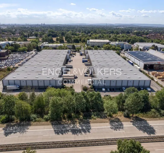 Aerial view of two large, gray commercial buildings with flat roofs, surrounded by trees and a highway. "Engel & Volkers Commercial" is displayed.