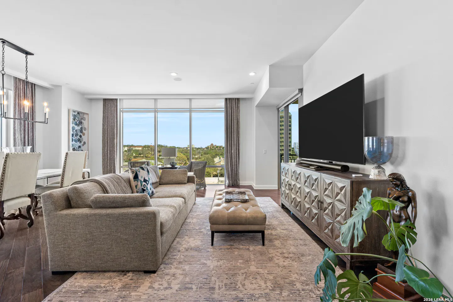 Bright living room with a gray sofa, ottoman, and patterned rug. A large TV sits above a decorative cabinet. Large windows offer a scenic view.