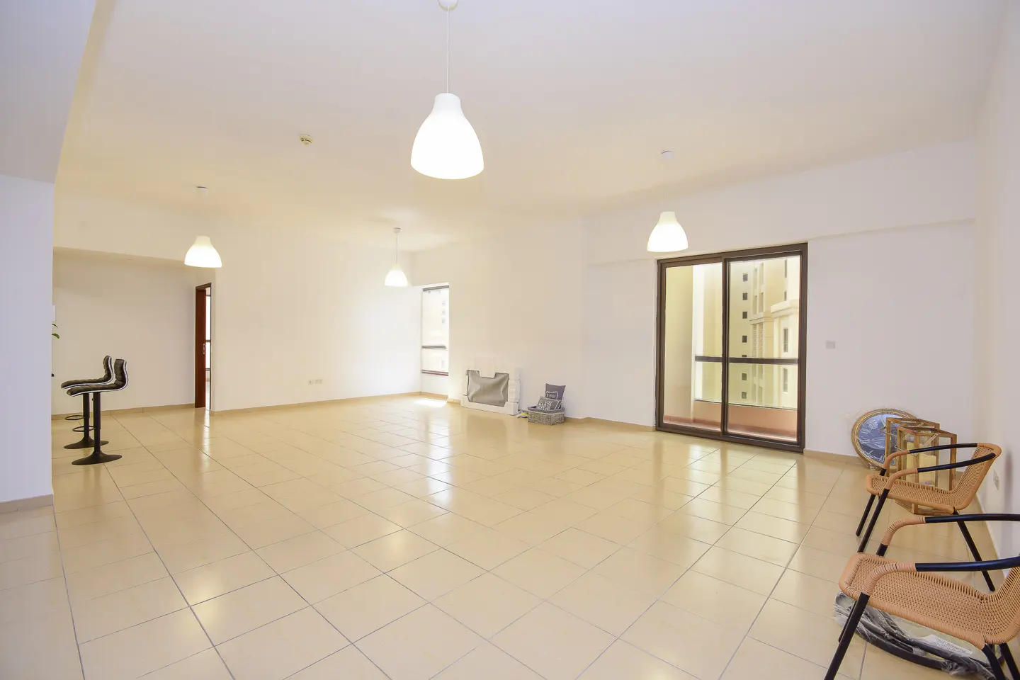 Bright, empty room with beige tile floor, white walls, and pendant lights. Chairs and bar stools are visible. A sliding glass door shows a building outside.