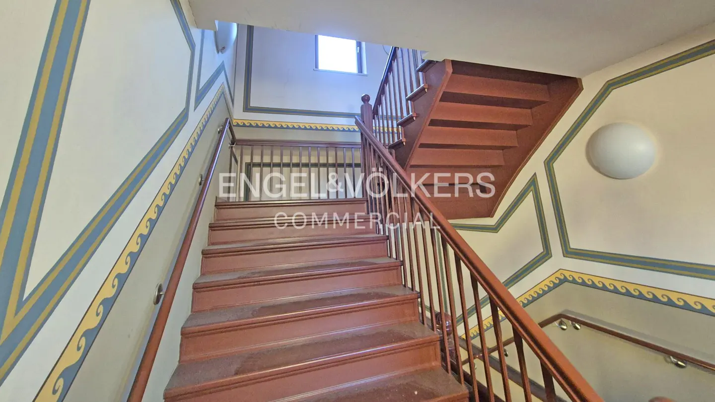 Interior view of a wooden staircase with brown handrails and decorative wall paintings in blue and yellow.