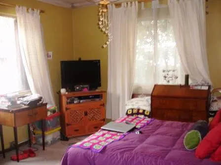 Bedroom with a purple comforter, wooden dresser, and TV. White curtains hang over a window. A laptop sits on the bed.