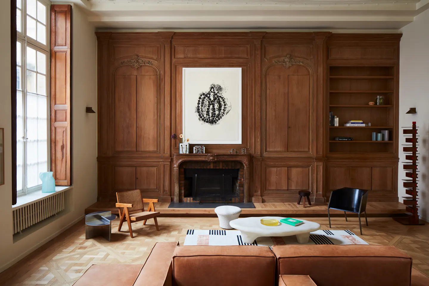 Living room with wood-paneled walls, a fireplace, abstract art, modern furniture, and a tan leather sofa on parquet floors.