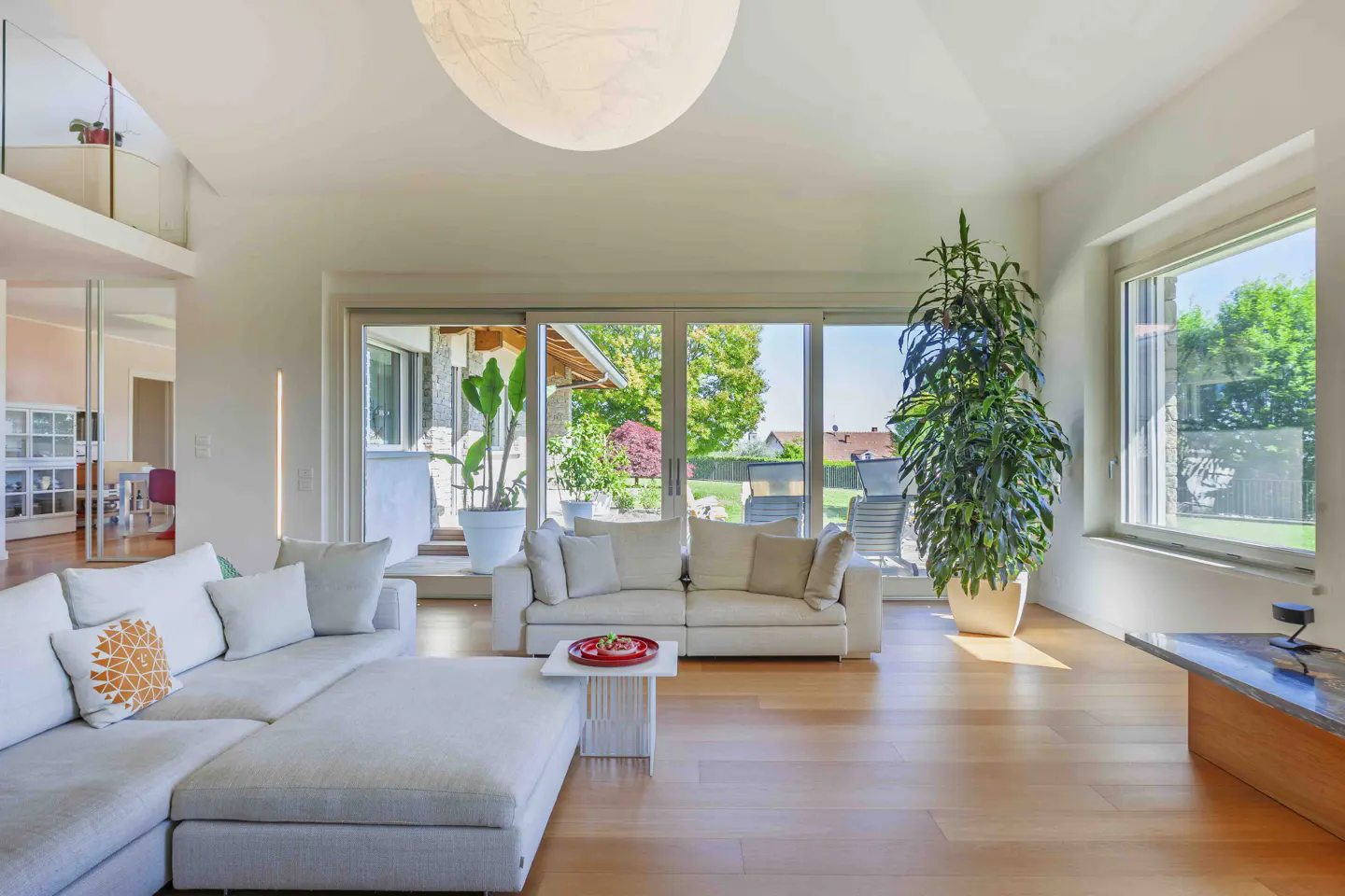 Bright living room with light wood floors, white sofas, and large windows overlooking a green yard. A large plant sits near the window.