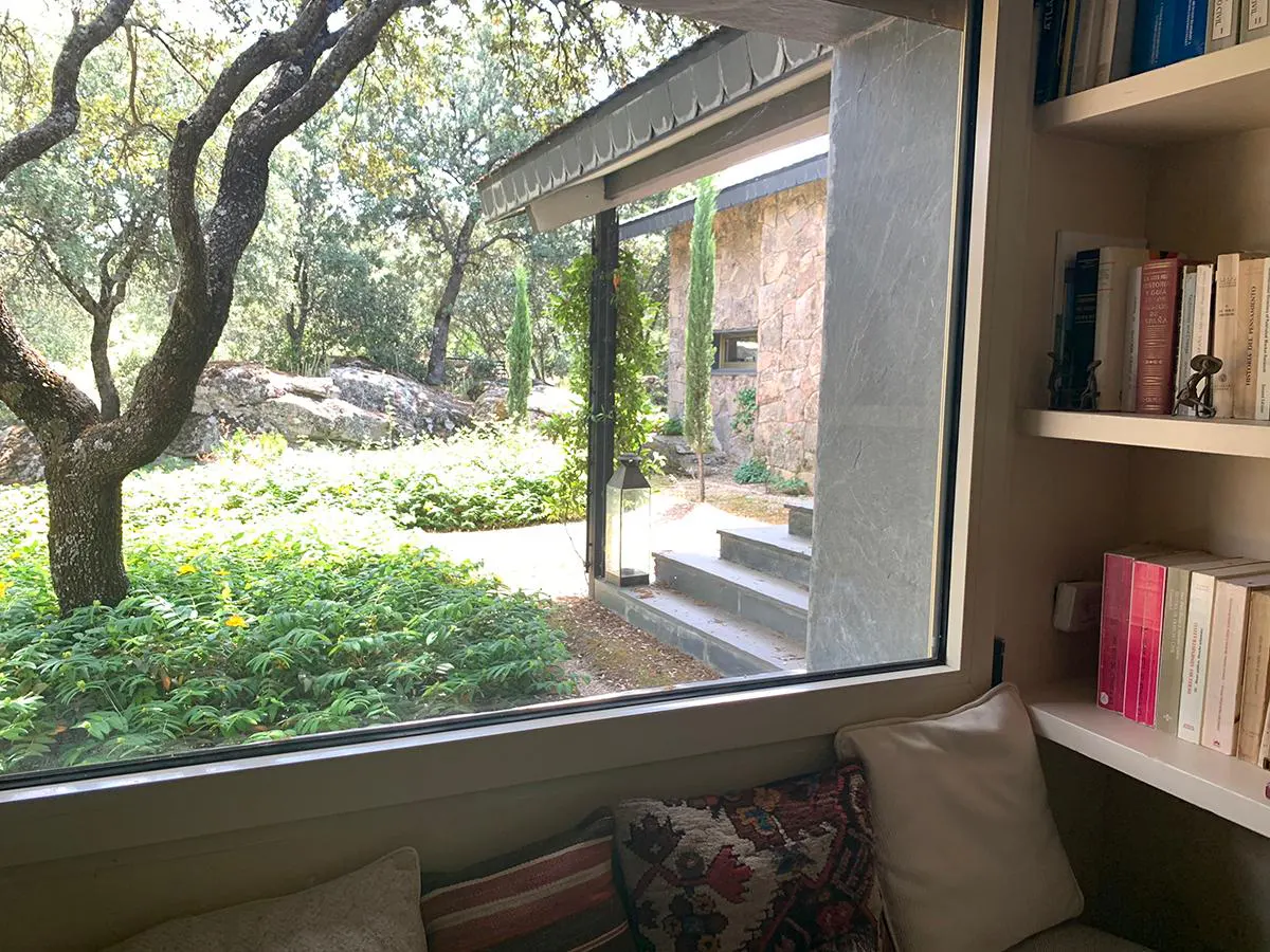 View from a window seat with pillows, looking out to a stone house, trees, and green plants. Bookshelves are on the right.