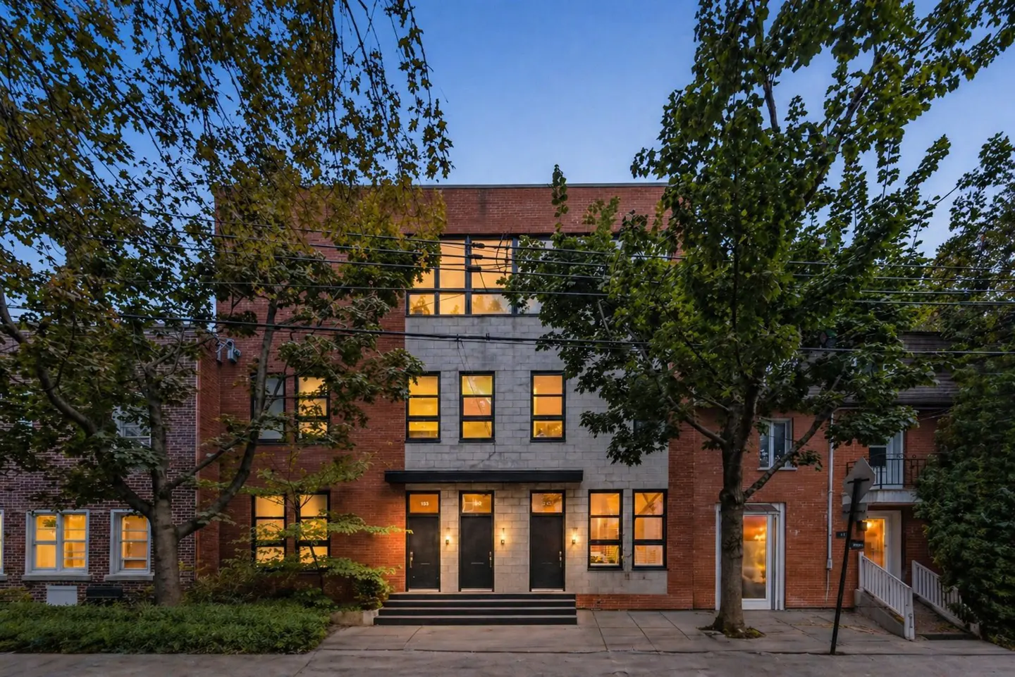 Exterior view of a three-story brick townhouse with black doors and window frames at dusk. Trees frame the building.