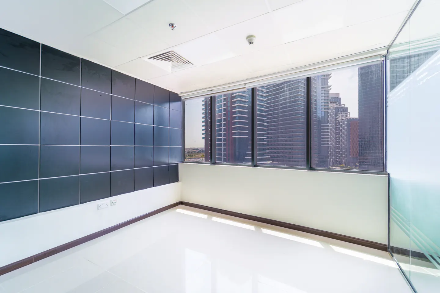 Empty office space with black tiled wall, white floor, and large window view of city skyscrapers.