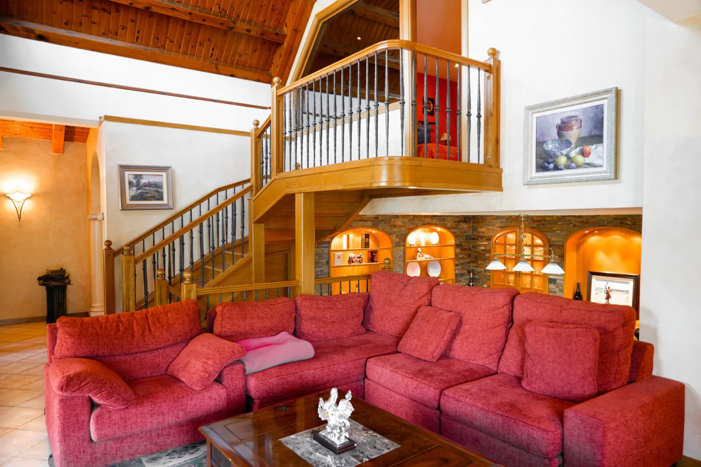 Living room with a red sectional sofa, wooden staircase, and a balcony with metal railings. The room has a high ceiling with wooden beams.