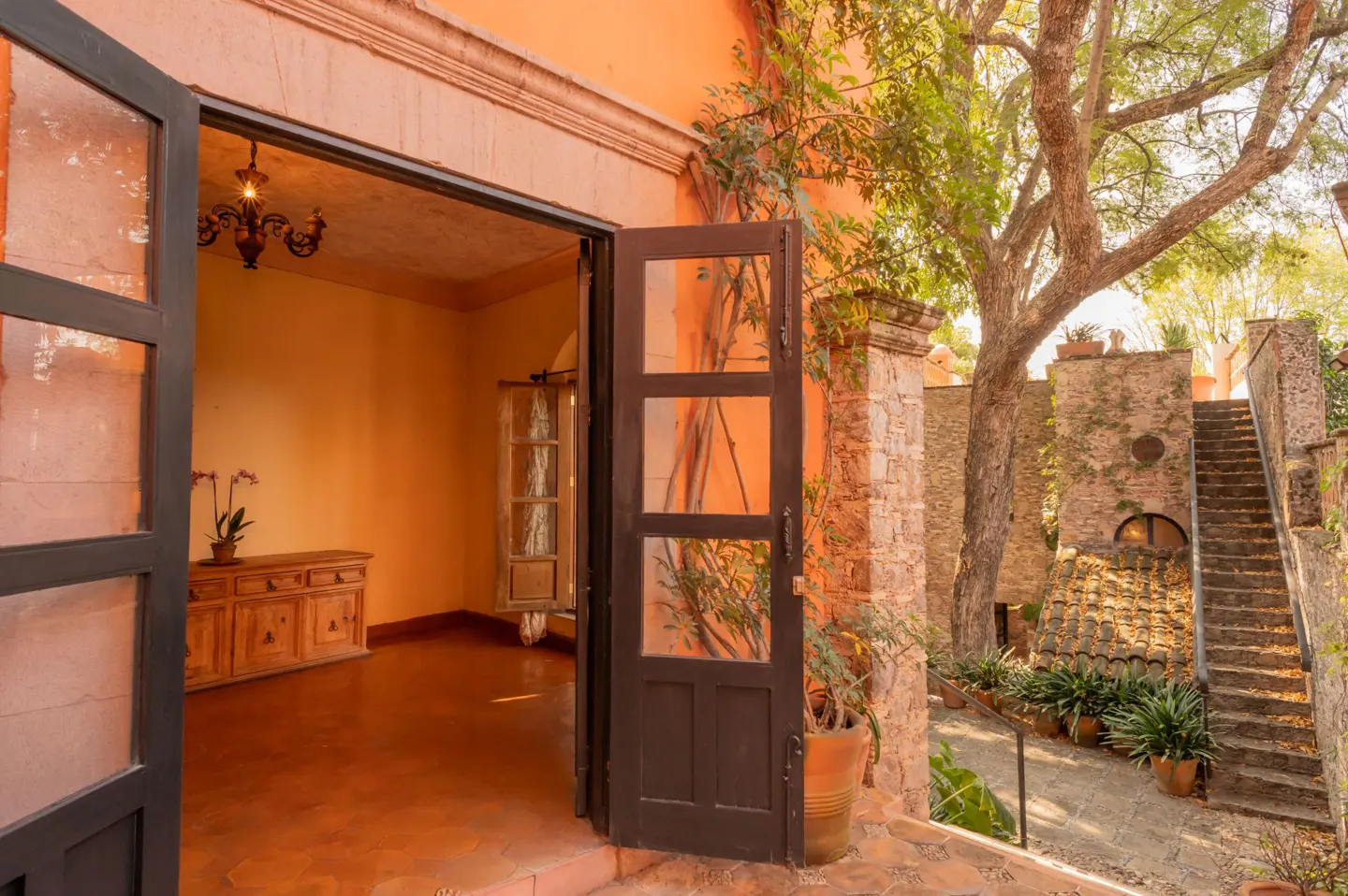 Open doorway of an orange building leads to a room with a chandelier and dresser, and a stone staircase outside.