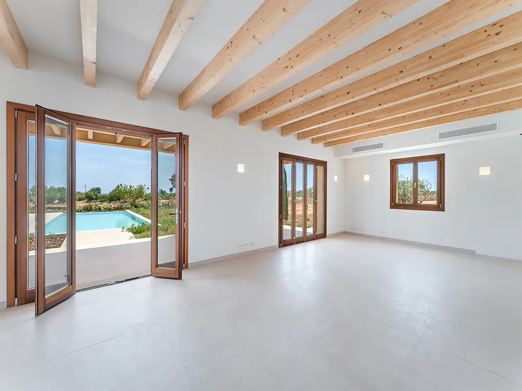 Bright, empty room with white walls, wood ceiling beams, and folding doors opening to a pool.
