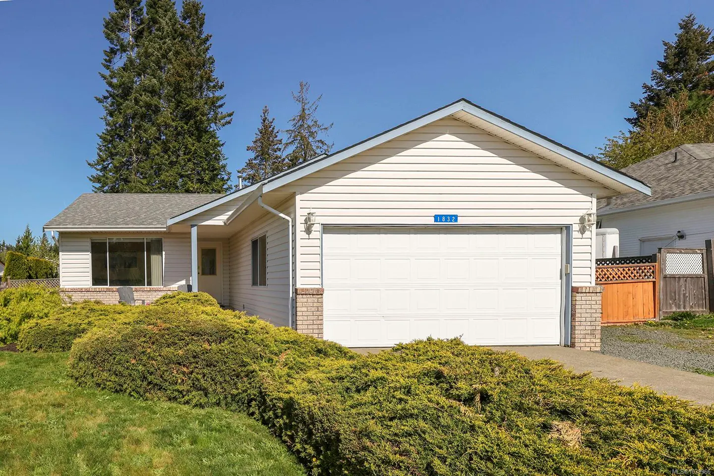 A single-story house with white siding, a white garage door, and green bushes in the front yard.