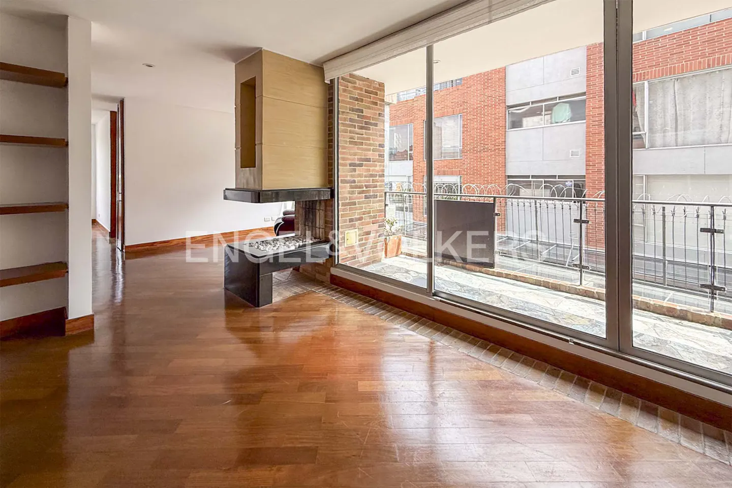 Bright living room with hardwood floors, a modern fireplace, and a large window overlooking a balcony and city buildings.