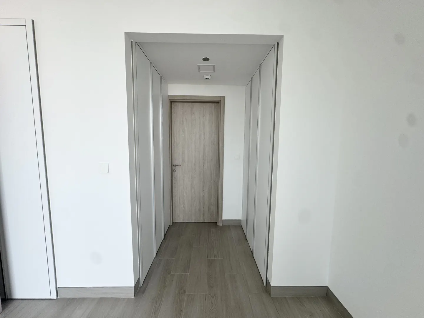 Interior view of a hallway with white walls, light wood floors, and a closed light wood door at the end.