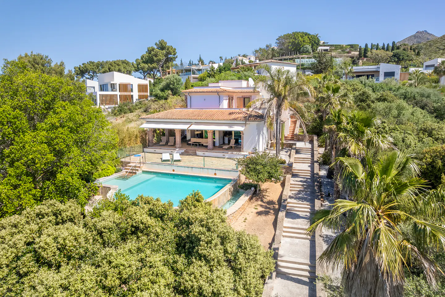 Aerial view of a white villa with a red tile roof, a blue pool, and stone steps surrounded by lush green trees.