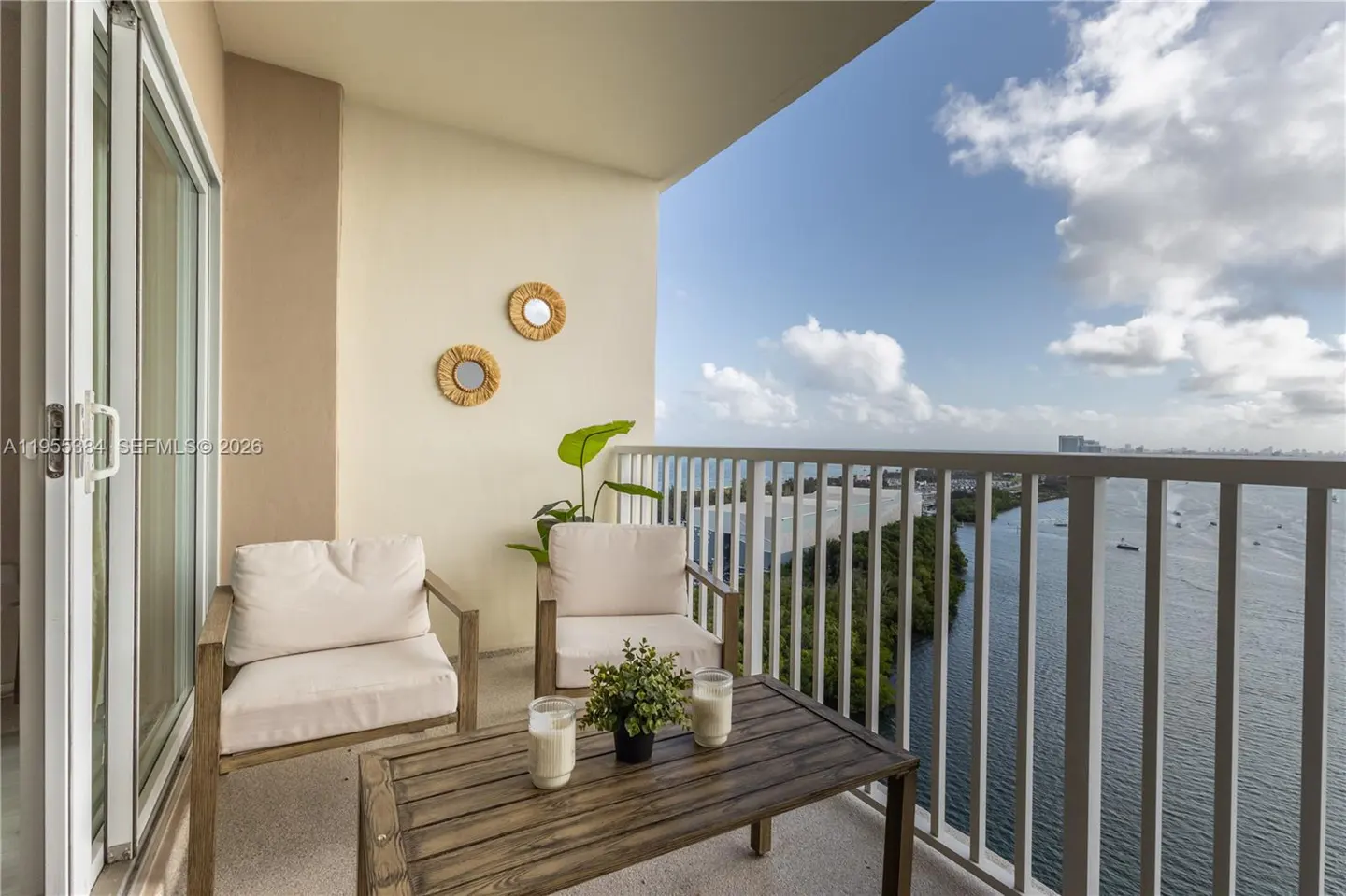 Balcony with two chairs, a wooden table, and a view of the water and sky. Two round mirrors hang on the wall.