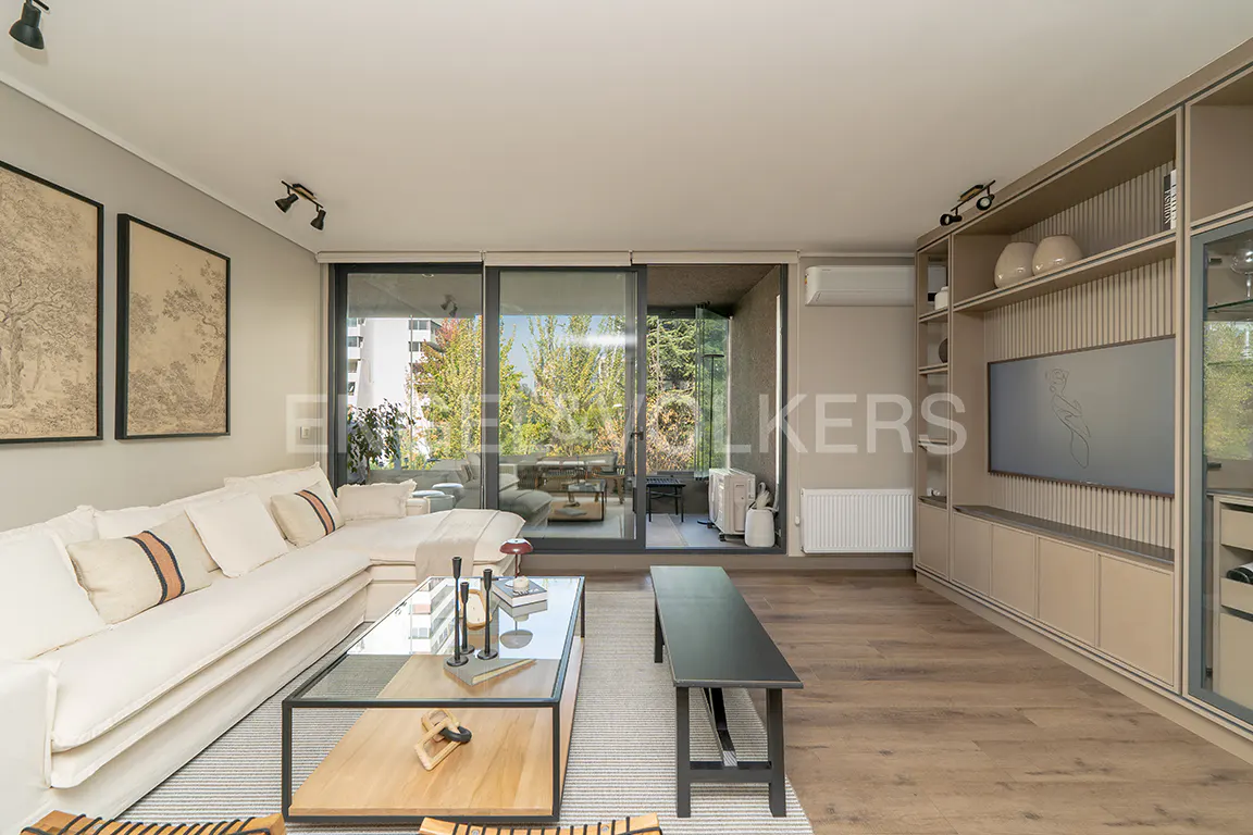 Living room with a white sectional sofa, glass coffee table, and a black bench. Sliding glass doors lead to a balcony with green trees. Built-in shelves and a TV are on the right.