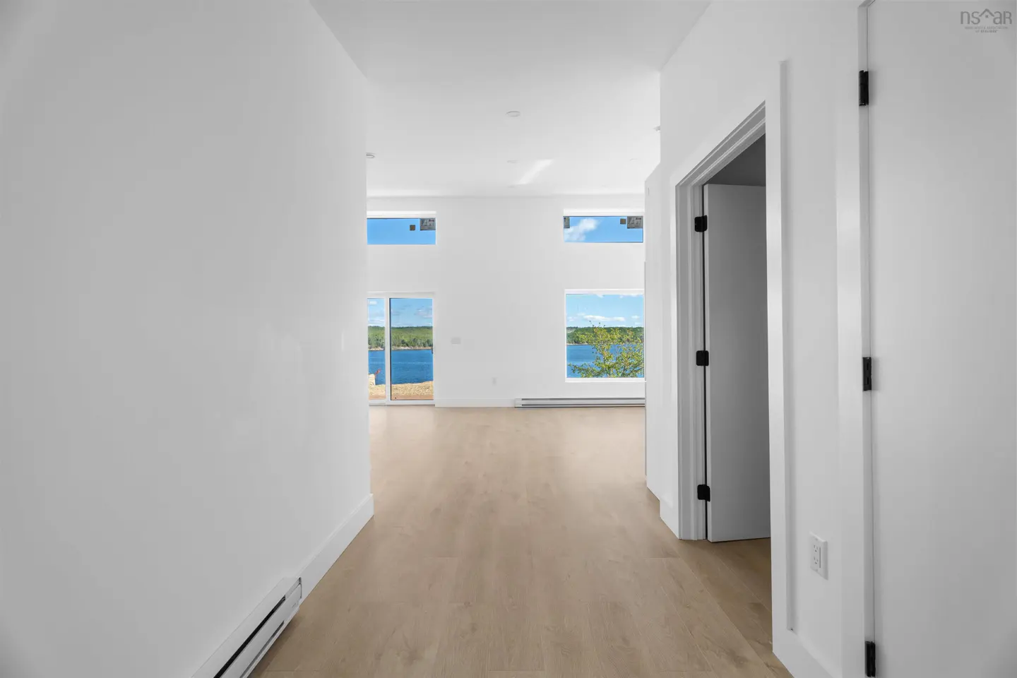 Bright, modern hallway with light wood floors and white walls. Large windows offer a scenic view of a blue lake and green trees.