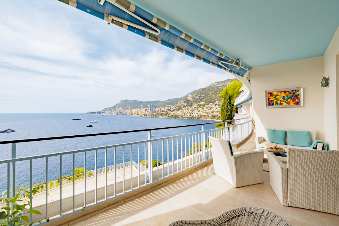 Spacious balcony with white wicker furniture, blue awning, and a panoramic view of the Mediterranean Sea and coastline. [AI-generated text]