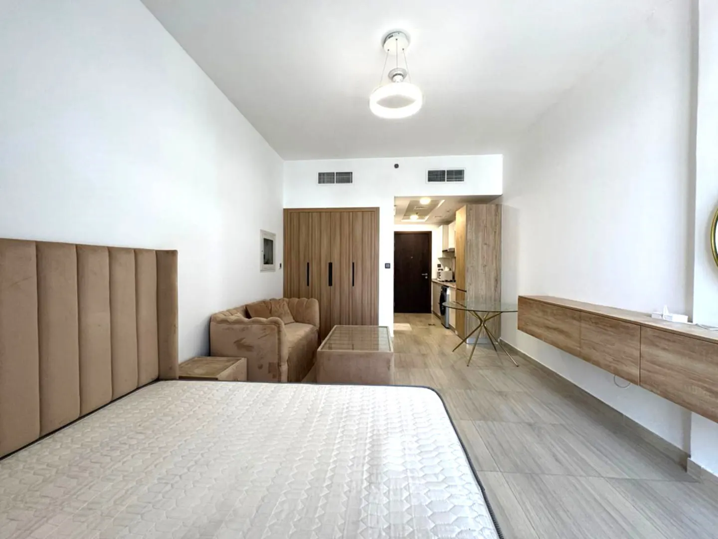 Studio apartment with a bed, sofa, and kitchen. The walls are white, and the floor is light gray. There is a light fixture on the ceiling.