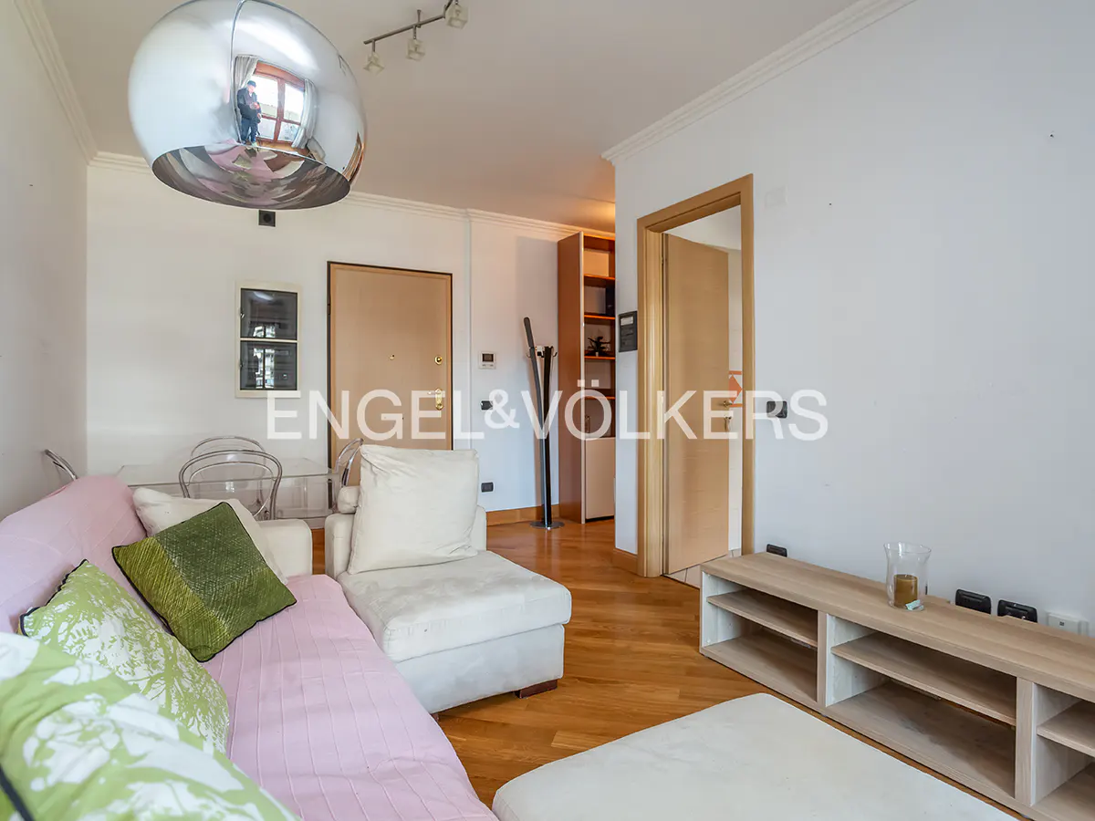 A bright living room with a pink sofa, green pillows, and a silver pendant lamp. A wooden floor and a light-colored TV stand are also visible.