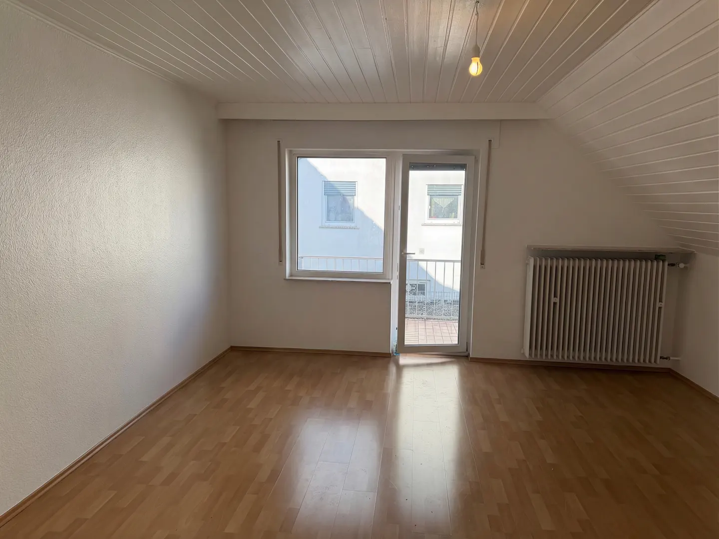 Bright, empty room with wood floors, white walls, and a white paneled ceiling. A window and door lead to a balcony. A white radiator sits on the right wall.
