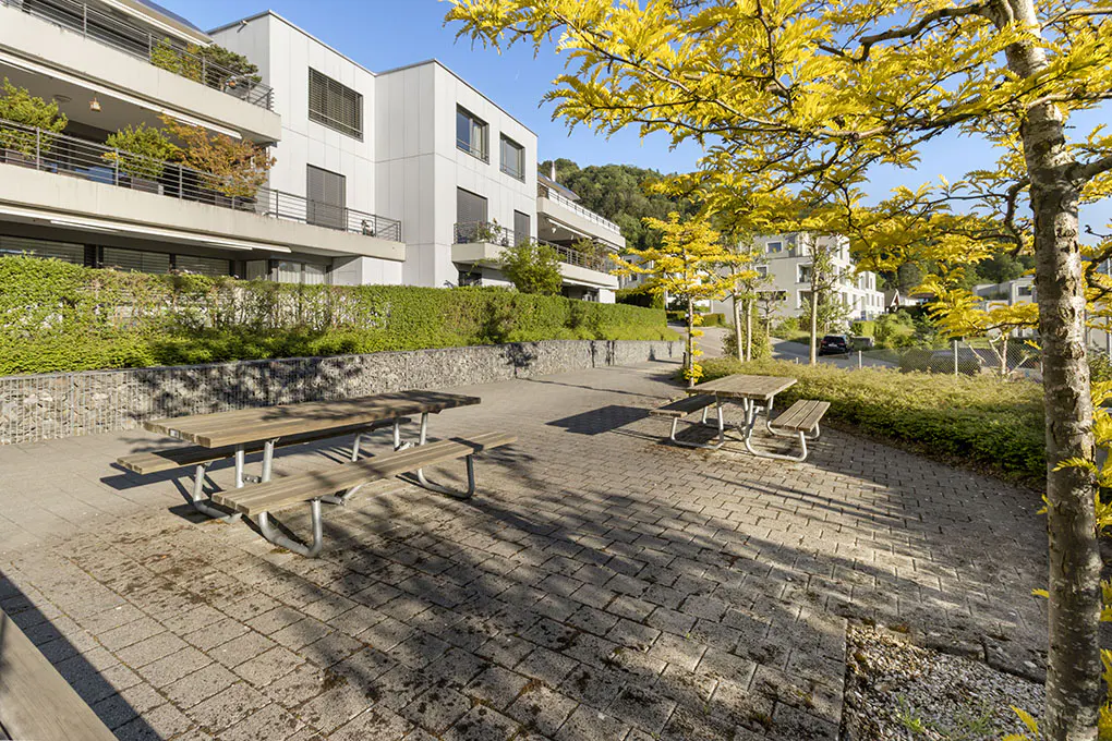 Outdoor patio with picnic tables in front of modern apartments. Yellow leaves on trees, green hedges, and stone walls.
