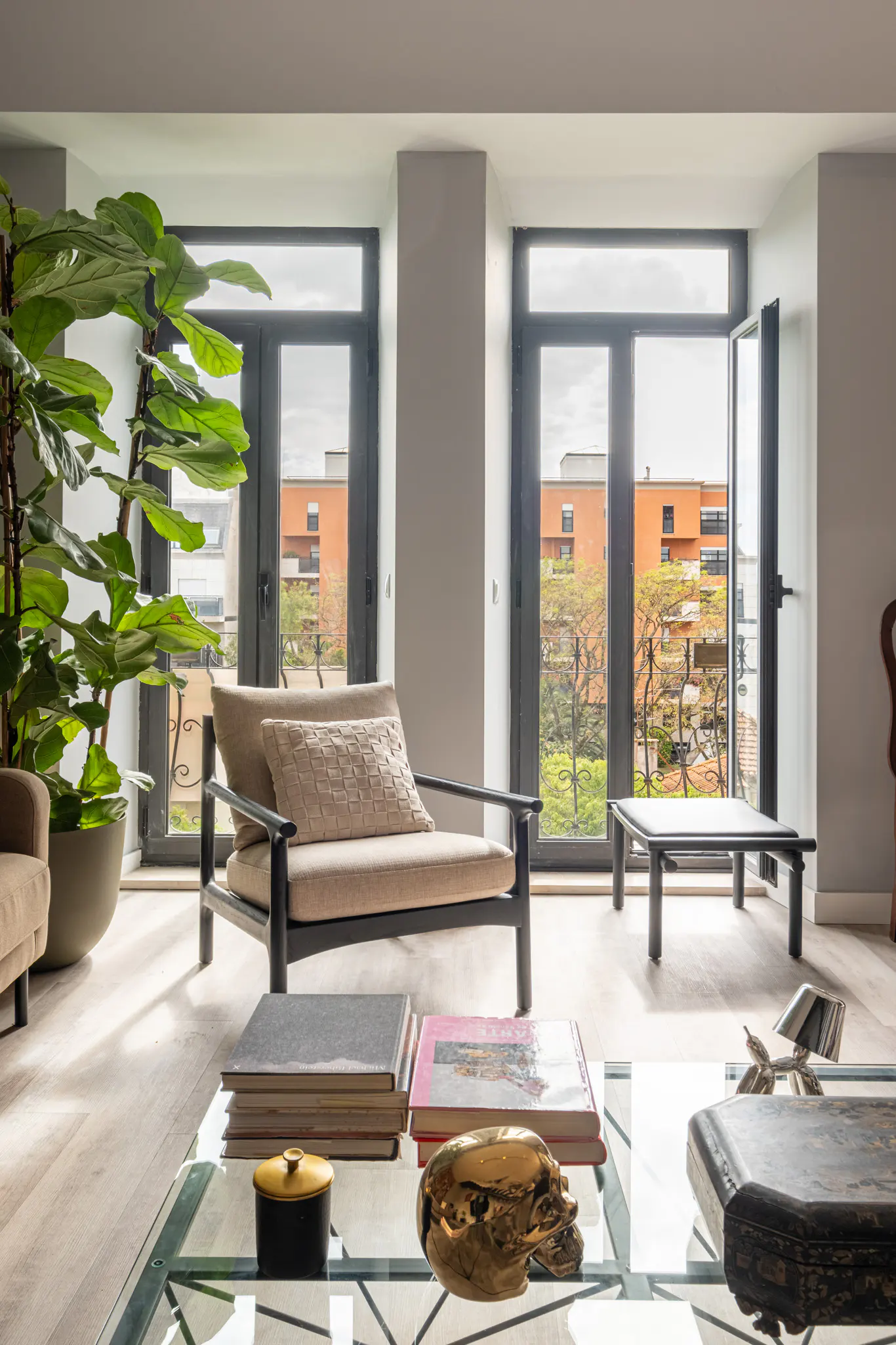 Bright living room with beige chairs, a glass coffee table with books, and a large potted plant near black-framed windows overlooking a city view.