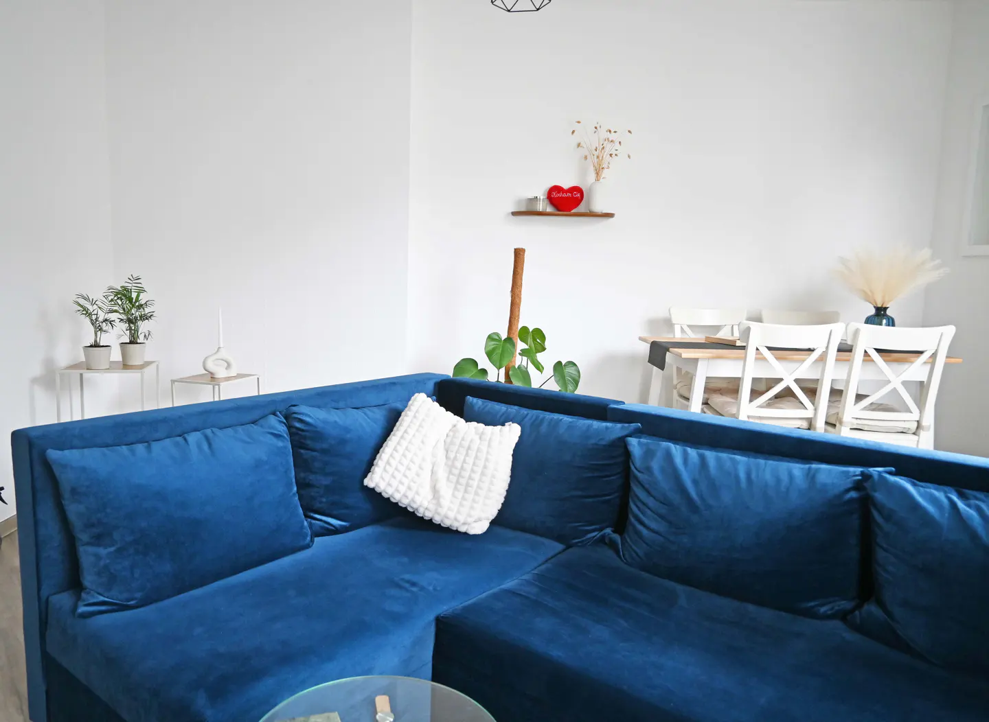 Bright living room with a blue velvet sectional sofa, white walls, and a dining table with white chairs in the background.