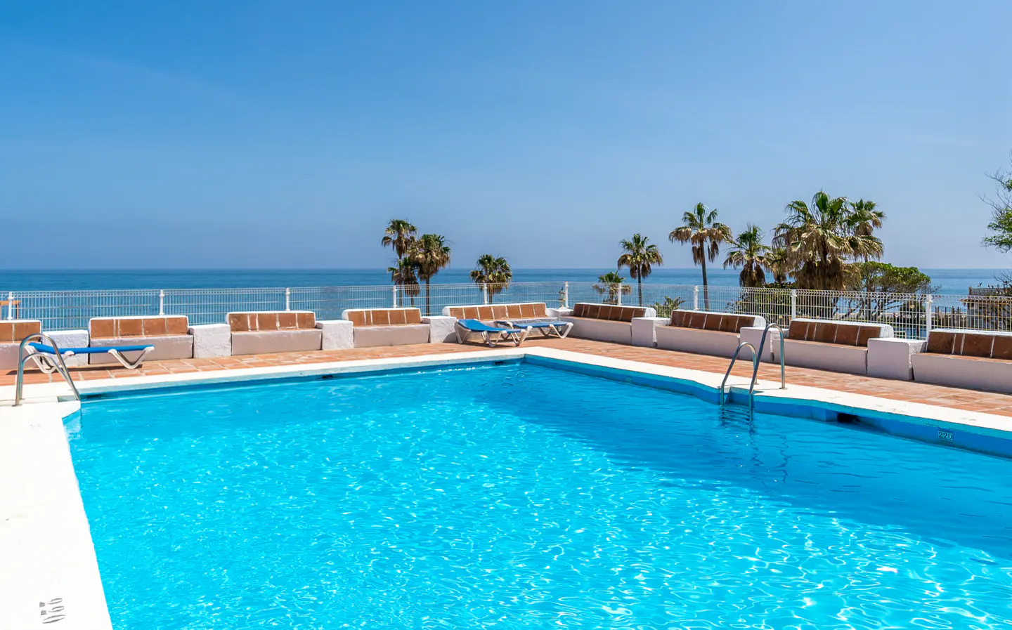 Outdoor pool with turquoise water, surrounded by white benches and lounge chairs. Ocean and palm trees in the background under a clear blue sky.