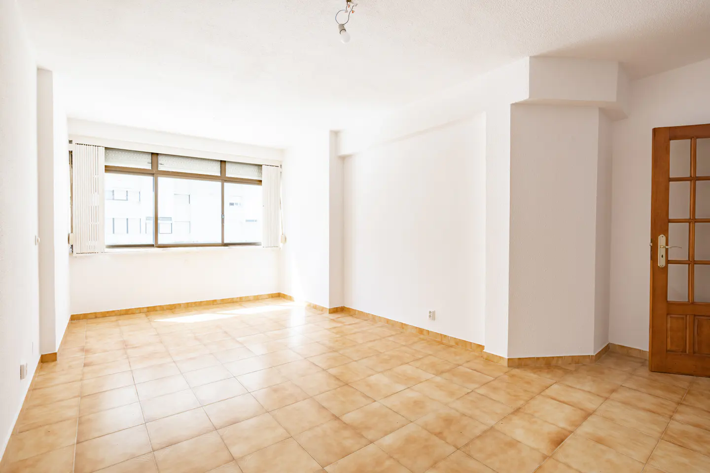 Bright, empty room with white walls, tan tile floor, and a large window with blinds. A wooden door with glass panes is on the right.